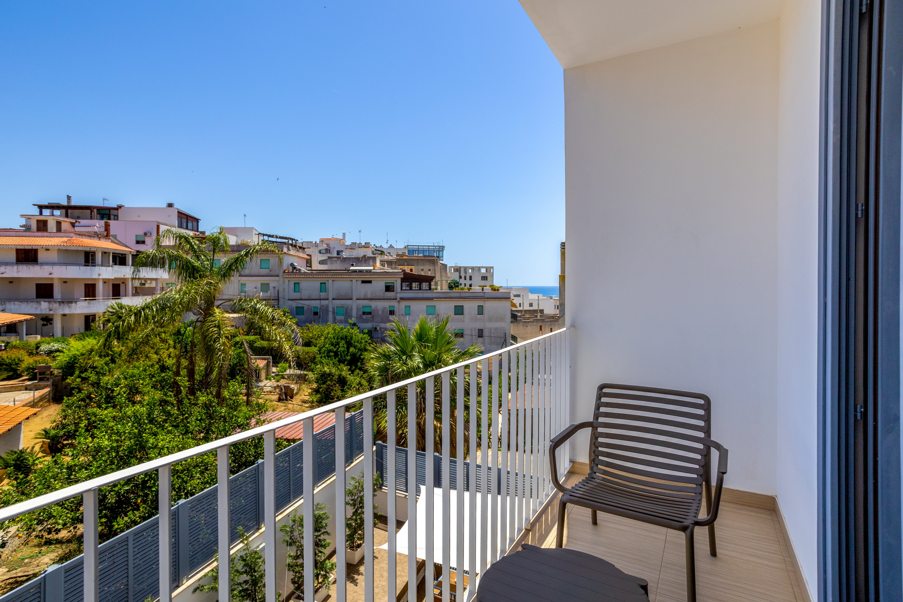 A small balcony with a modern chair and table overlooks a coastal town under a clear blue sky. Residential buildings and lush green palm trees fill the middle ground, with a glimpse of the sea visible in the distance. The bright sunlight and clean white railings create a warm, inviting atmosphere. The scene suggests a relaxing outdoor space in a vacation apartment or hotel.