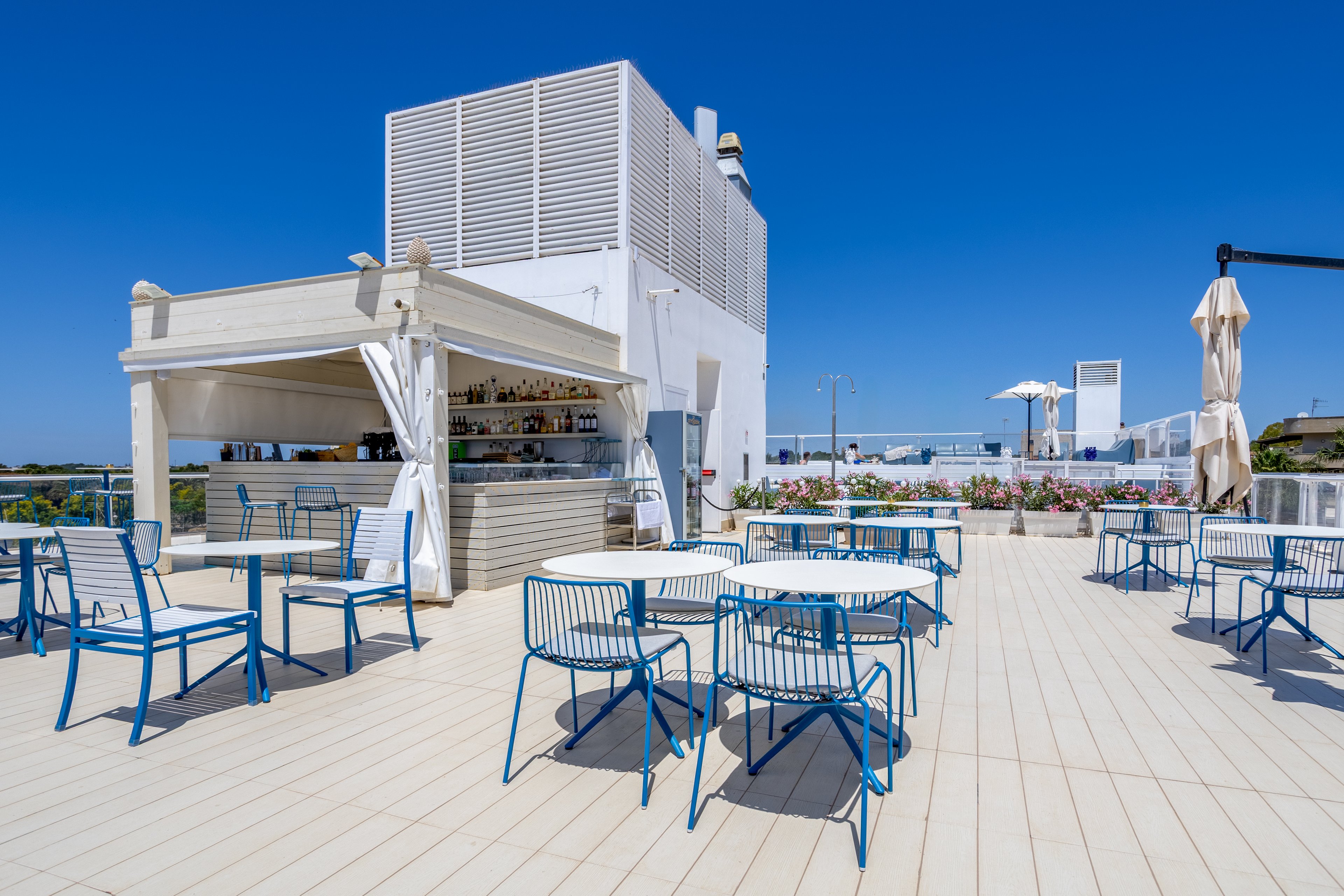 An open-air rooftop bar features modern blue and white tables and chairs under a clear blue sky. A white bar kiosk stocked with bottles stands to the side, with a few umbrellas and planters adding detail to the bright terrace. The spacious deck has light-colored flooring and offers an unobstructed view of the surrounding area. The scene feels calm, inviting, and ideal for outdoor drinks or relaxation.