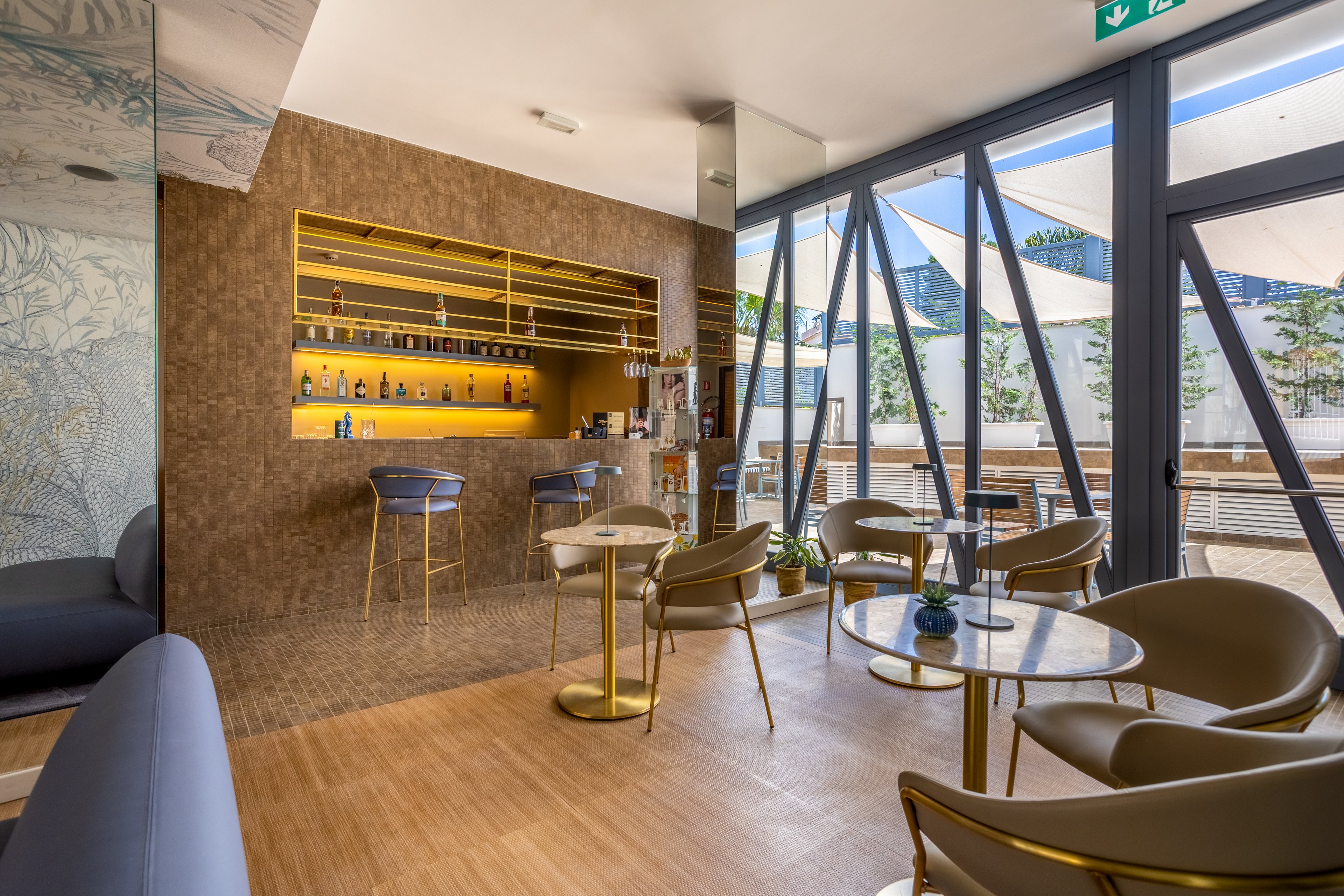 Contemporary bar lounge interior featuring a small counter with bottles displayed on golden shelves. Stylish chairs and round tables are arranged on a light wooden floor, creating an inviting seating area. Large glass windows with geometric frames open onto a terrace with umbrellas and mountain scenery. The overall atmosphere is bright and modern, with warm metallic accents and natural light.