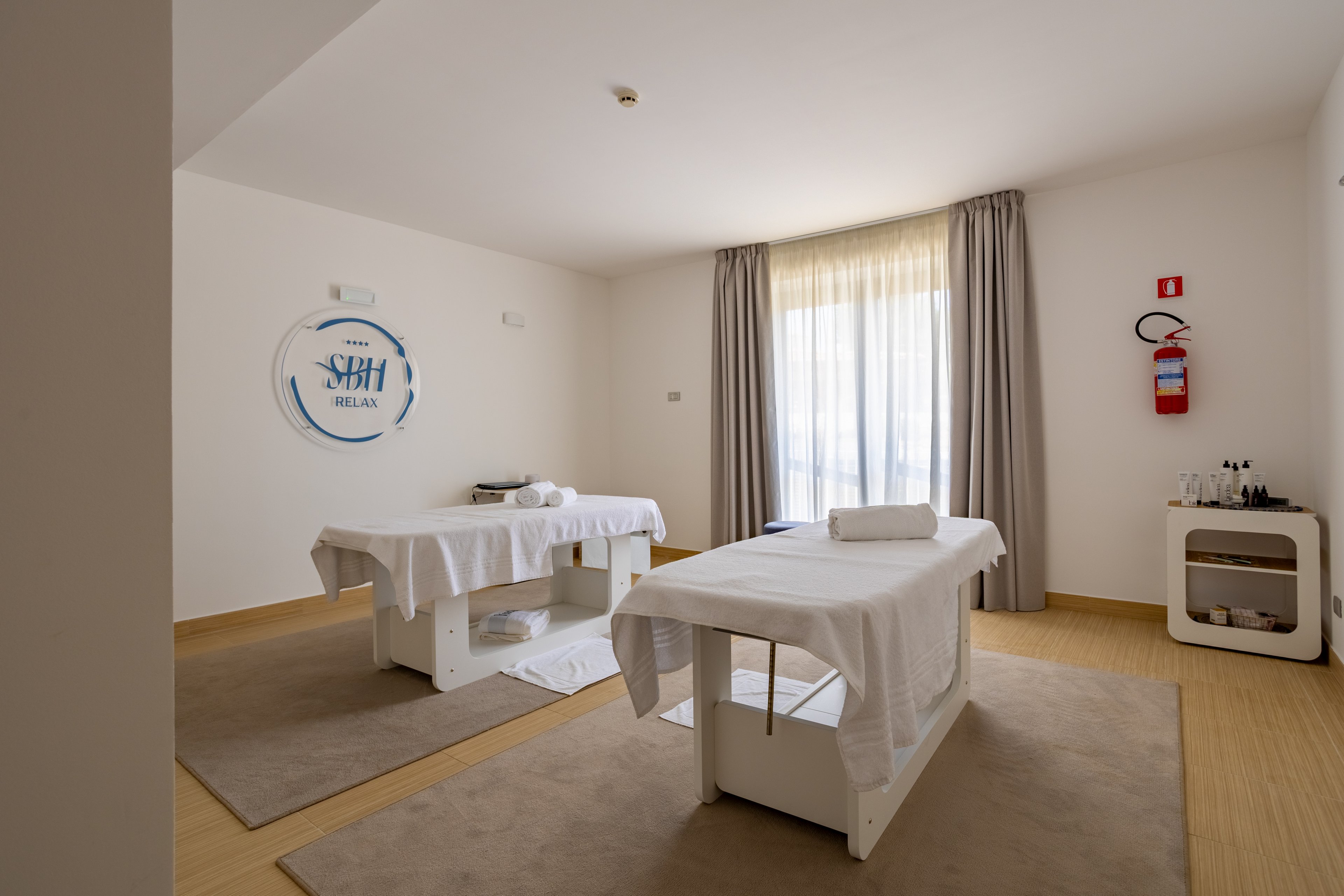A serene spa treatment room features two massage tables neatly prepared with white linens and towels. Natural light filters through a large window with sheer curtains, creating a calm and inviting atmosphere. Neutral walls and beige flooring enhance the minimalist, clean design. A wall-mounted fire extinguisher and a small station with bottles and supplies are visible, adding functional details to the space.