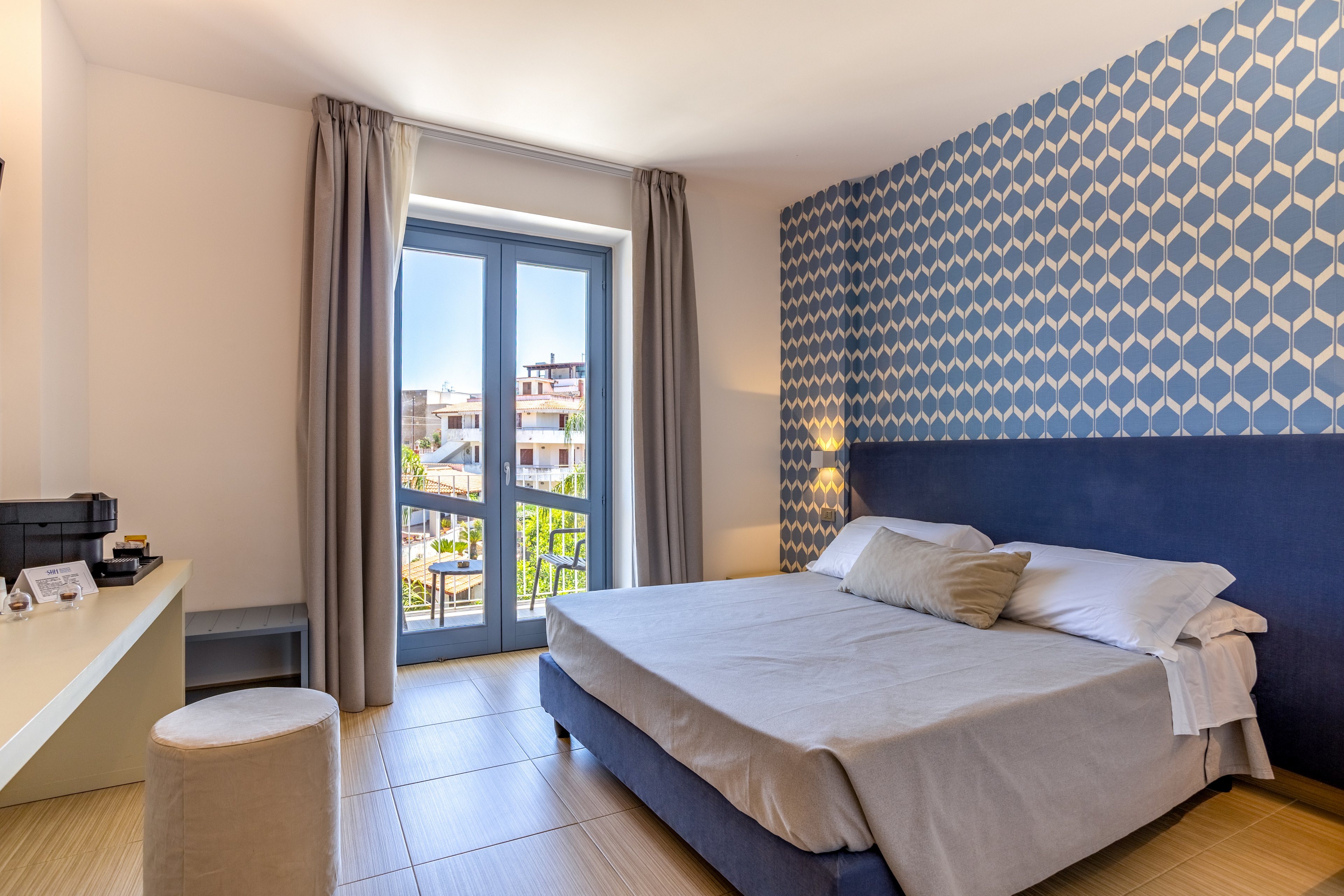 Contemporary double bedroom with a large upholstered bed and neutral bedding. The room features patterned blue accent wallpaper, light wood flooring, and floor-to-ceiling curtains framing glass balcony doors. A small desk area with coffee machine and accessories sits opposite the bed, alongside a round ottoman. Sunlit buildings and greenery are visible through the balcony doors, adding an airy, inviting feel.