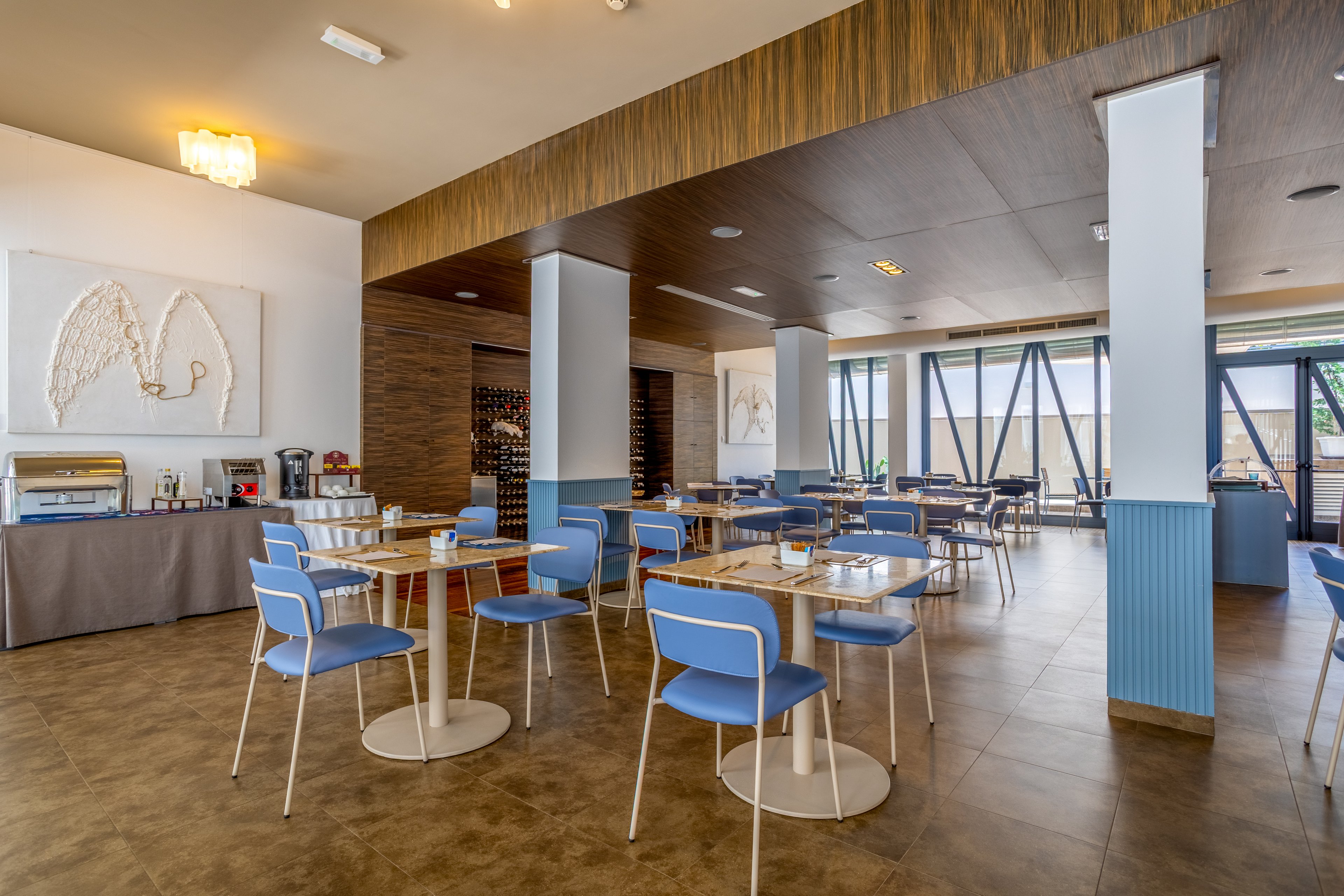 A contemporary cafeteria interior features blue chairs and round white tables arranged neatly in rows. Large windows on one side allow natural light to brighten the space, while wood and pastel accents create a warm, inviting atmosphere. A buffet counter with food service equipment is visible along the wall, and decorative artwork hangs above it. The room appears clean, organized, and ready to welcome guests.