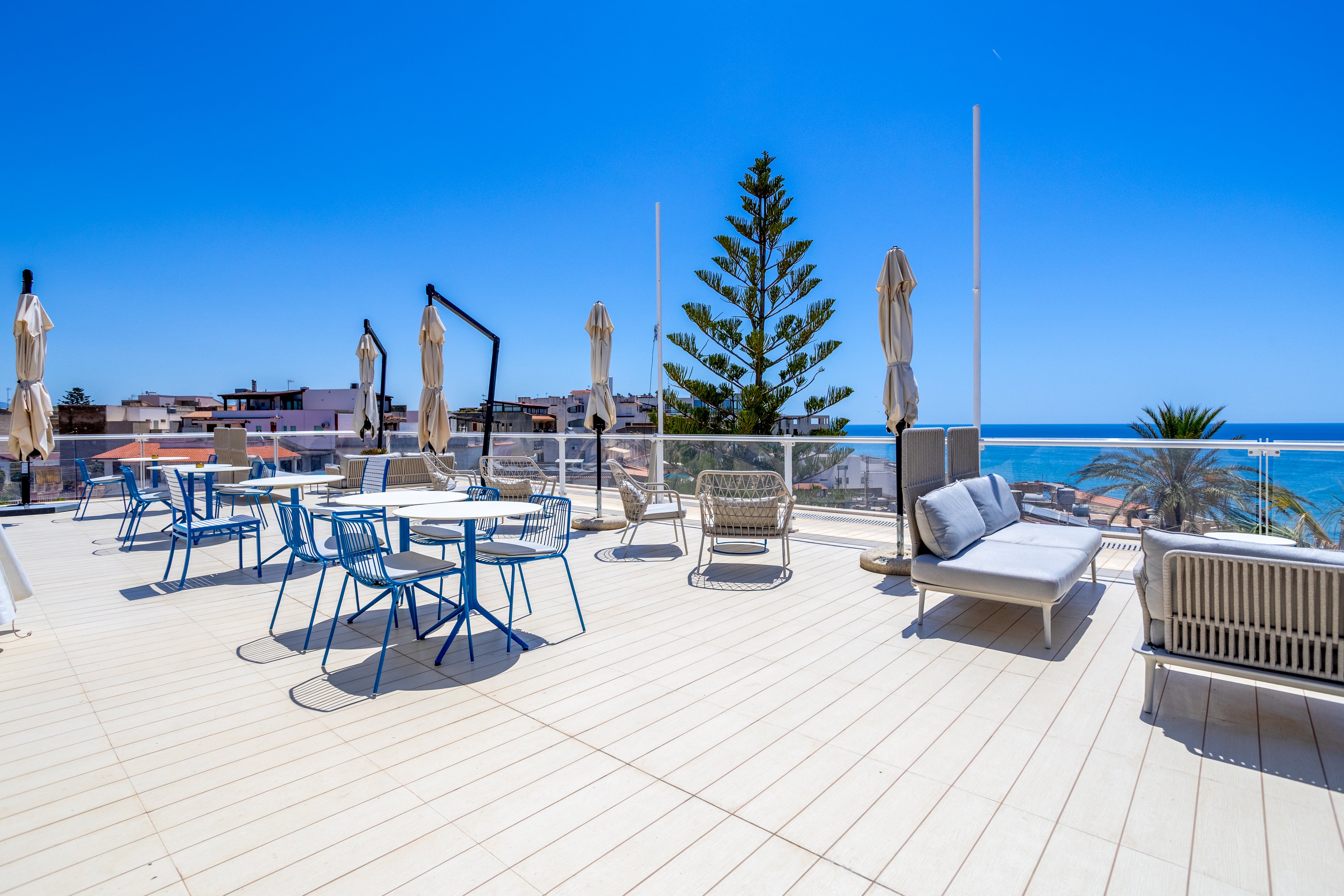 An open rooftop terrace features modern outdoor seating and tables under a bright blue sky. Closed umbrellas stand around the deck, suggesting a relaxed leisure or dining area. In the background, coastal buildings and a tall evergreen tree frame the view. The ocean is clearly visible, adding a serene, vacation-like atmosphere to the scene.