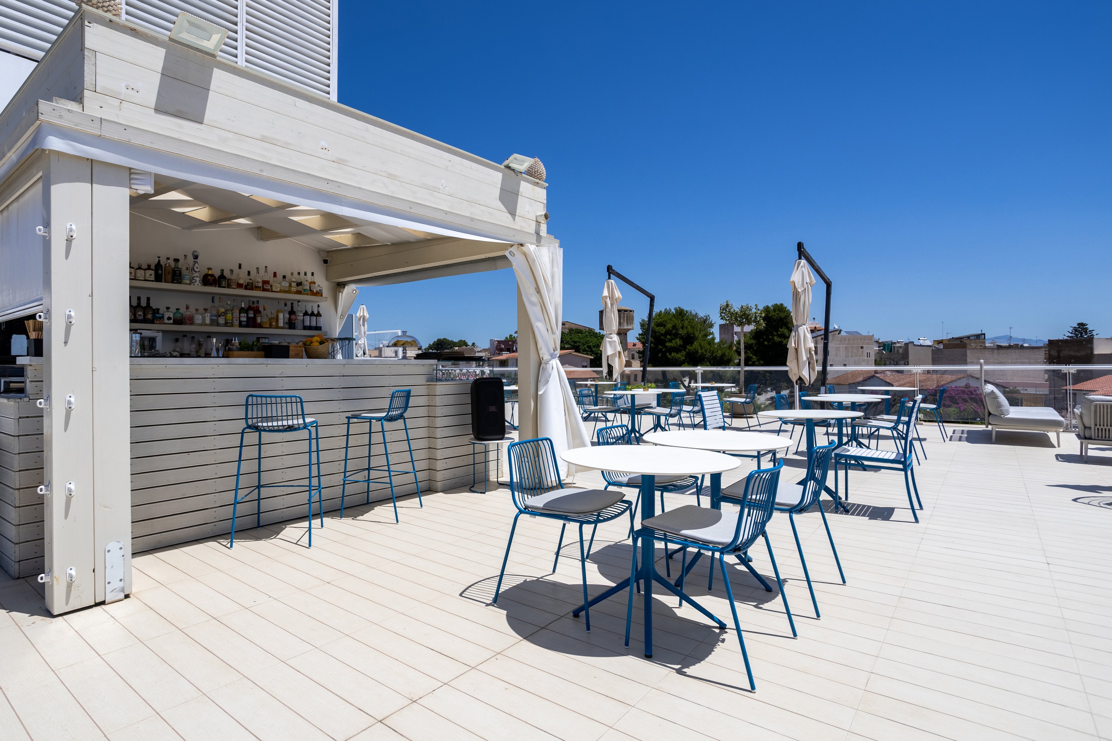 An open-air rooftop bar features a light-colored counter with bottles and glassware neatly arranged behind it. Blue metal barstools and matching chairs surround round tables on a spacious deck. Large umbrellas and white structural elements provide shade under a clear blue sky. Nearby buildings and trees are visible in the background, suggesting an urban setting.