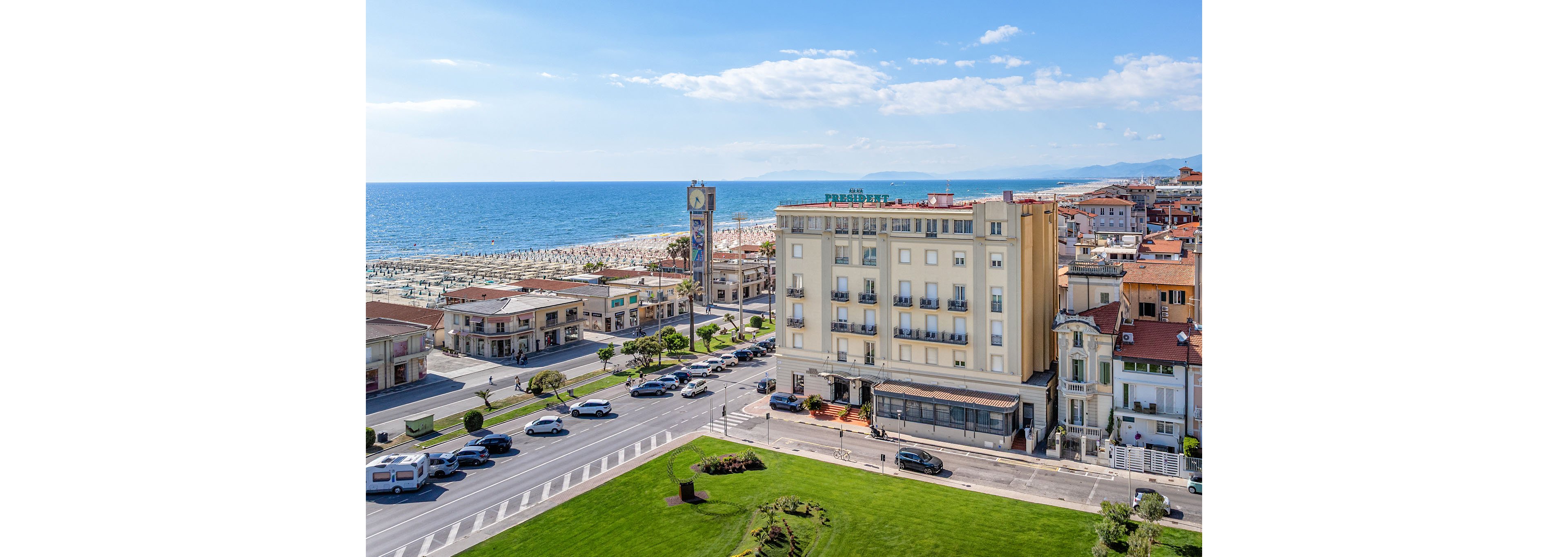 An elevated view shows a seaside town with a large multi-story hotel facing the waterfront. A broad road and green lawn with landscaped flowerbeds run in front of the building, while cars travel along the coastal promenade. Beyond the town, rows of beach umbrellas line the sandy shore next to the calm blue sea. A tall clock tower stands near the beach, and distant hills are visible under a bright, partly cloudy sky.