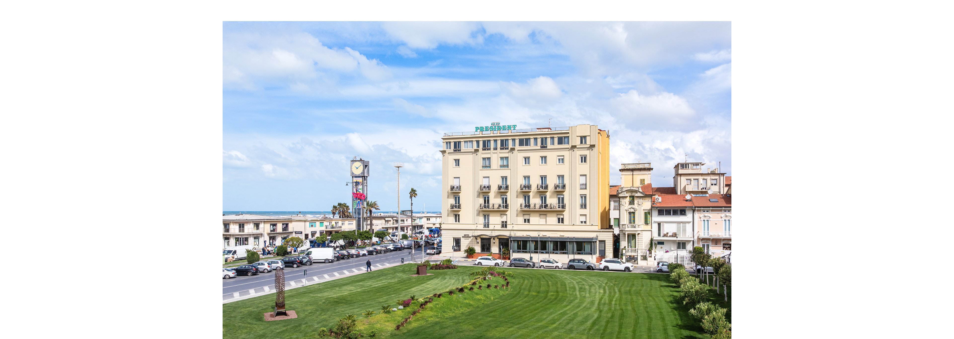 A mid-rise hotel building stands prominently along a coastal city street. In the foreground, a neatly manicured green lawn with landscaped shrubs leads toward the busy road. Cars and pedestrians move along the waterfront avenue, with a clock tower and additional low-rise buildings visible in the background. The sea and cloudy blue sky frame the urban seaside setting, creating a bright and open atmosphere.