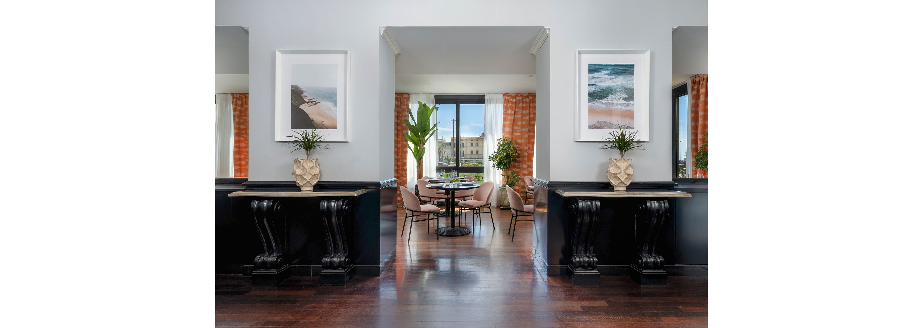 A stylish interior features a central dining area framed by two black console tables with sculptural bases and decorative vases. Soft pink chairs surround a round black table near a large window that looks out onto a bright building and greenery. Coastal-themed framed artwork hangs on the light gray walls above each console, while tall plants and patterned orange curtains add color and texture. The polished dark wood floor and abundant natural light create a clean, contemporary atmosphere.