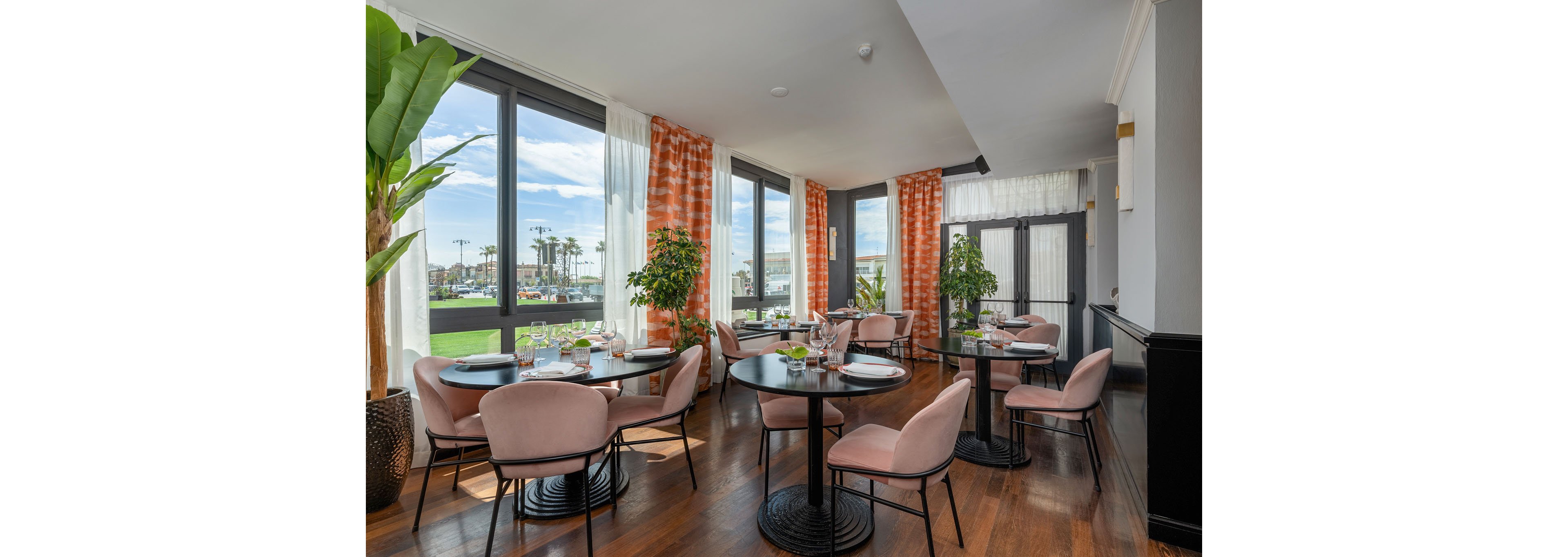 Contemporary restaurant interior features round black tables and soft pink upholstered chairs arranged neatly for guests. Large windows let in abundant natural light and offer a view of palm trees and an outdoor green space. Sheer white curtains with orange patterned drapes frame the windows, adding warmth and color to the room. Potted plants, set tables with glassware, and polished wooden floors create an inviting, upscale dining atmosphere.