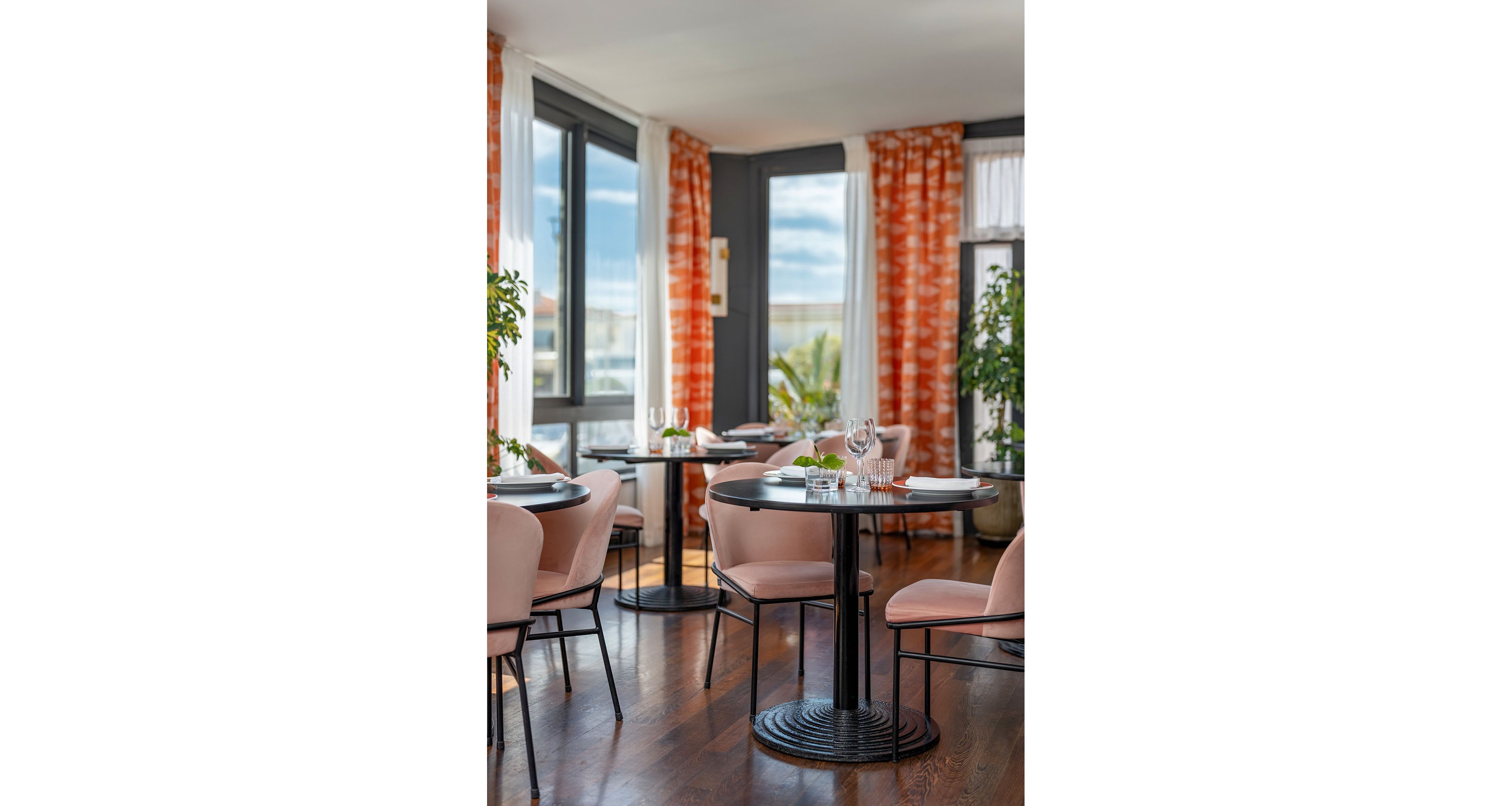 A modern restaurant dining area features round black tables set with plates, glasses, and cutlery. Soft pink upholstered chairs surround the tables on a polished wooden floor. Large windows let in natural light, framed by orange patterned curtains and white sheers. Green potted plants add a fresh touch to the airy, inviting space.