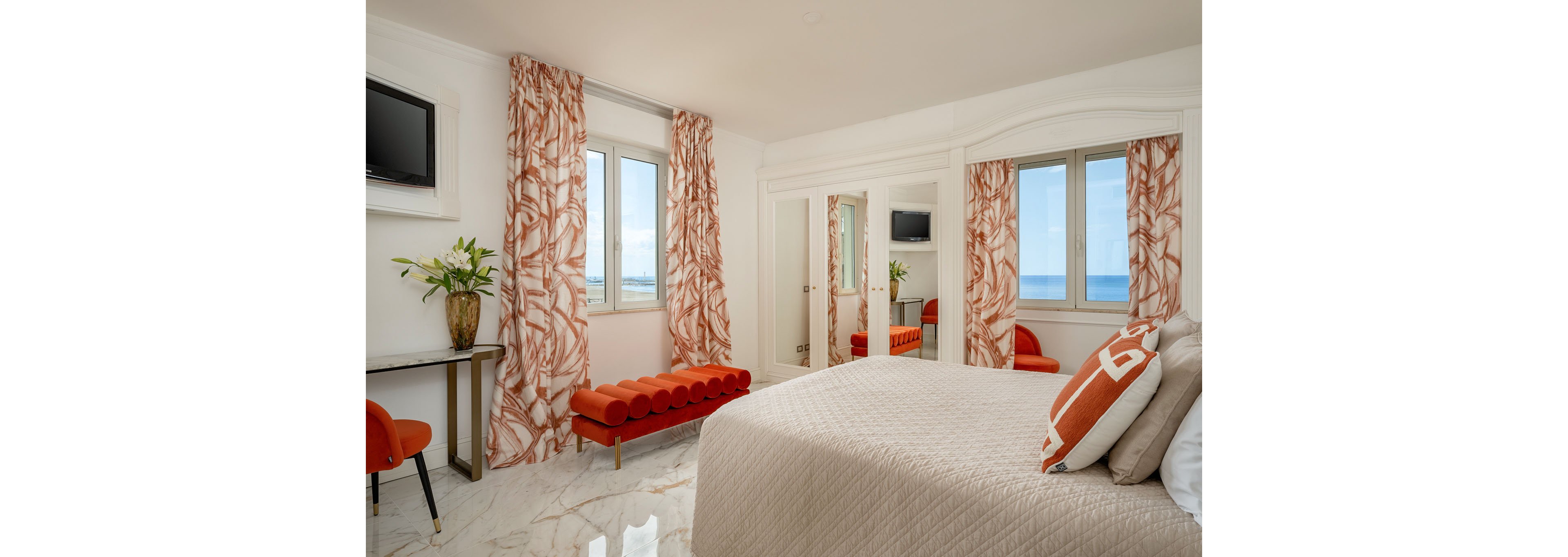 Elegant hotel bedroom featuring a neatly made bed with patterned accent pillows and a quilted bedspread. Large windows with patterned curtains frame a clear ocean view, filling the room with natural light. A red upholstered bench and matching chair add bold color against the white walls and marble floor. A wall-mounted television, mirrored closet doors, and a small desk with a vase of flowers complete the contemporary coastal decor.