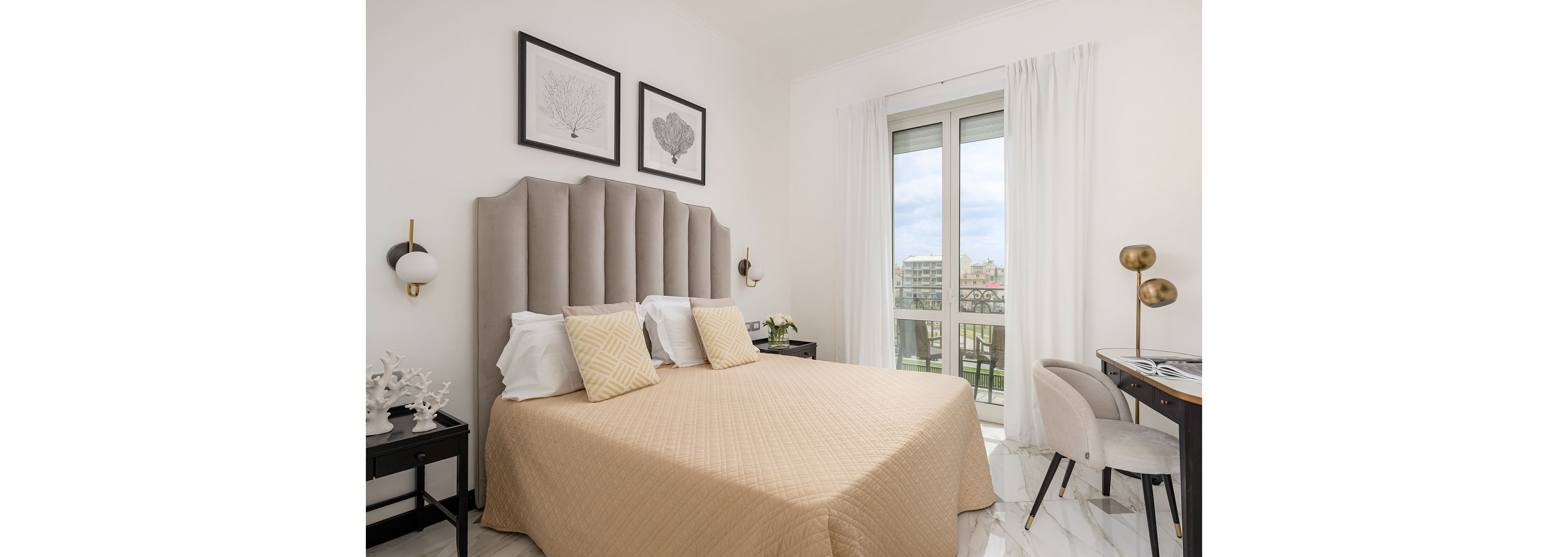 A contemporary bedroom features a large bed with a tall upholstered headboard and neutral bedding accented by patterned pillows. The room is bright, with white walls, marble-style flooring, and sheer curtains framing a glass door that opens to a balcony overlooking city buildings. Matching black bedside tables hold decorative objects and wall-mounted sconces, while a small desk and upholstered chair create a compact workspace. Overall, the space feels airy, minimalist, and elegantly styled.