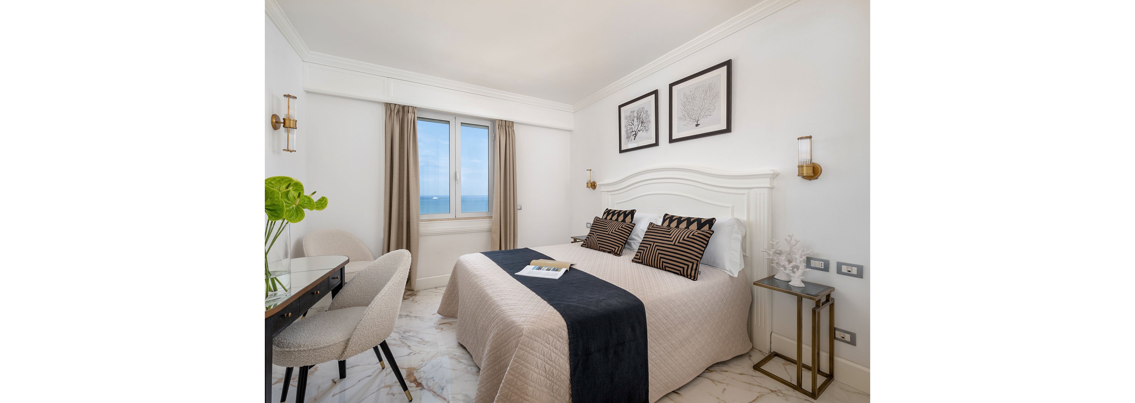 Elegant double bedroom with a large white headboard and two patterned pillows on a neatly made bed. A window with beige curtains reveals a clear blue ocean view, adding a coastal feel to the space. The room features marble flooring, a small desk with a chair, and a side table with decorative items. Wall-mounted lights and framed artwork above the bed contribute to the clean, contemporary style.