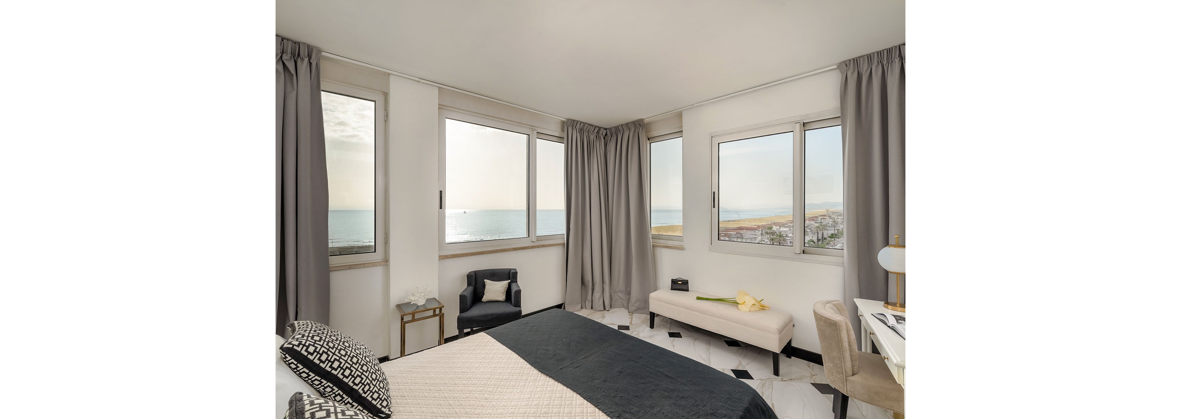 A modern bedroom features large windows overlooking a calm sea and coastline. The room includes a bed with dark bedding, a cushioned bench, and an upholstered armchair beside a small side table. Light curtains frame the expansive view, while neutral tones and minimal decor create an airy, relaxing atmosphere. Marble-style flooring and soft furnishings add a touch of contemporary elegance.