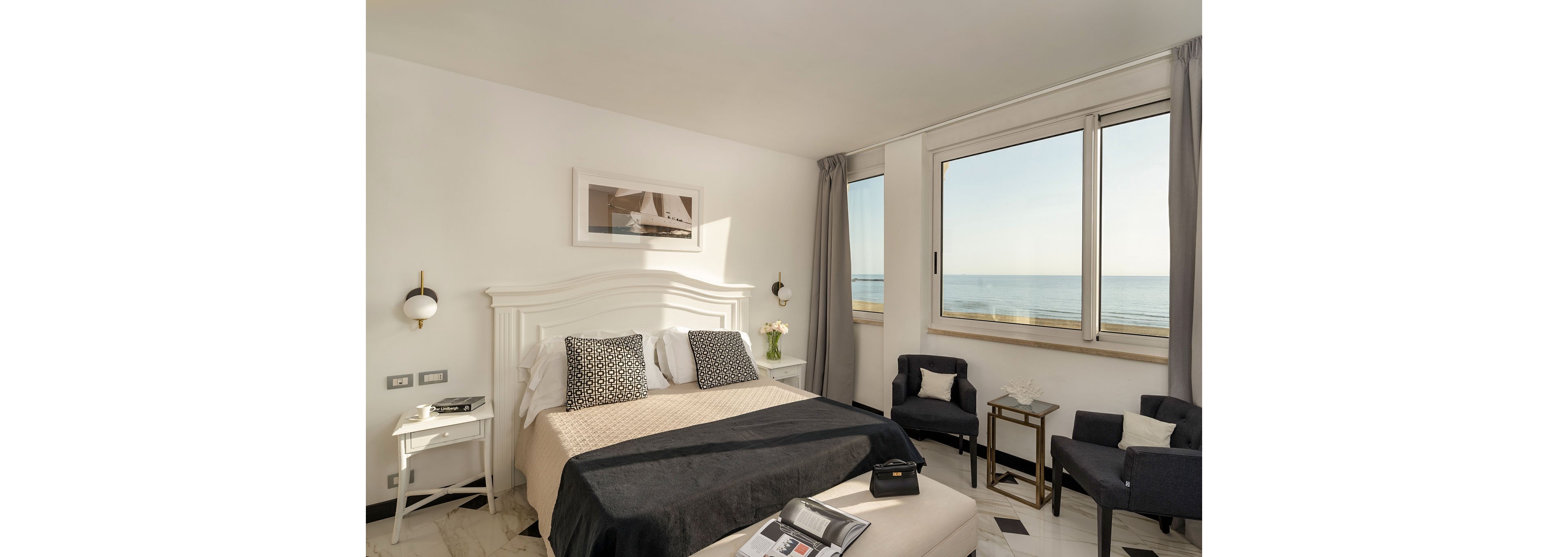 A neatly made double bed with neutral linens and patterned pillows sits in a bright hotel bedroom. Large windows reveal a calm ocean view, filling the room with natural light. Two dark armchairs, side tables, and a small ottoman create a cozy seating area. Minimalist decor, soft gray curtains, and a framed nautical photograph contribute to a clean, contemporary coastal style.