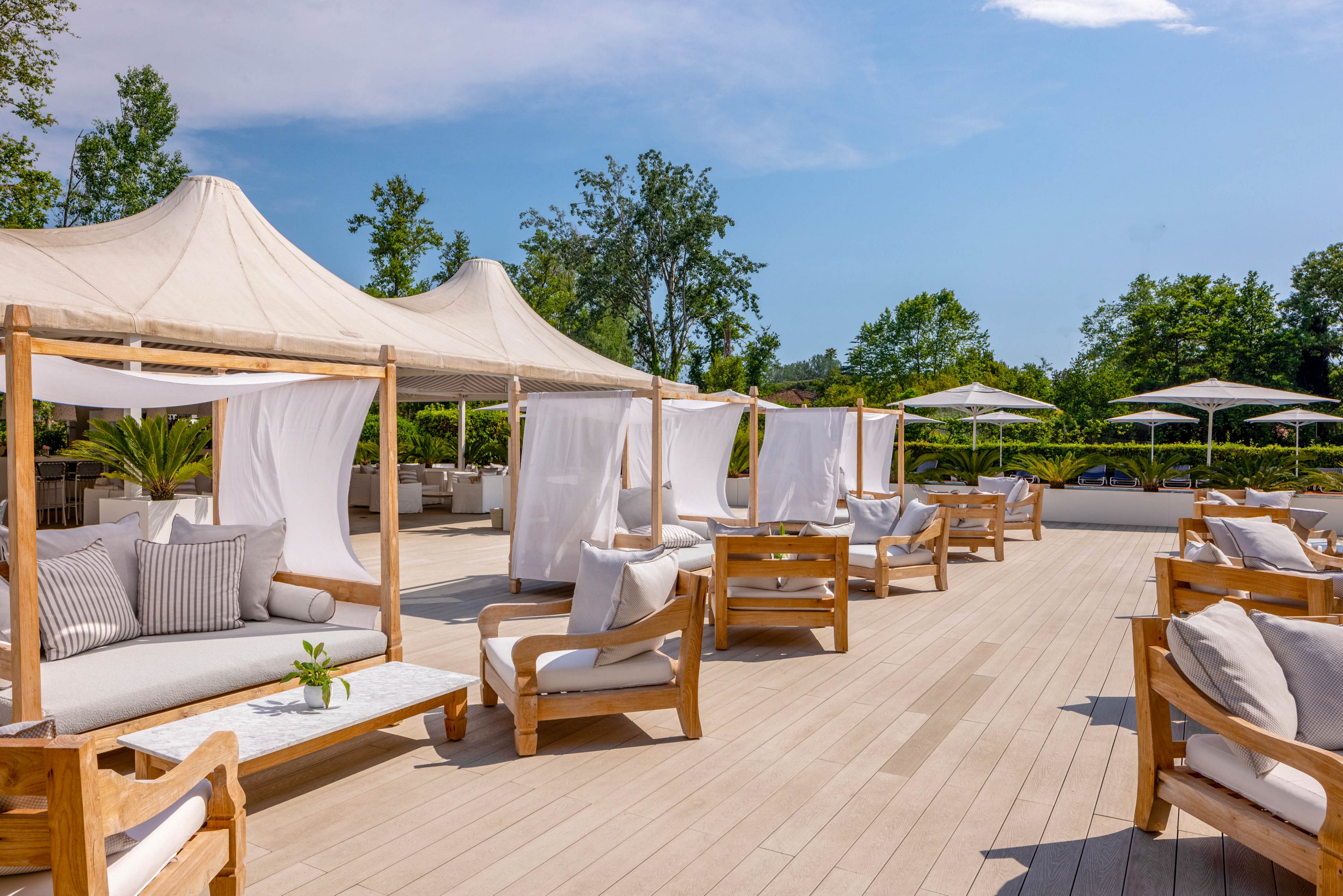 Spacious outdoor lounge features wooden daybeds with white canopies and plush cushions, arranged on a light wooden deck. The setting is open-air, surrounded by greenery and shaded by large fabric tents, creating a relaxed and inviting atmosphere. The scene is bright and airy, with neutral tones and natural materials enhancing the tranquil mood. No visible text or numbers are present in the image.
