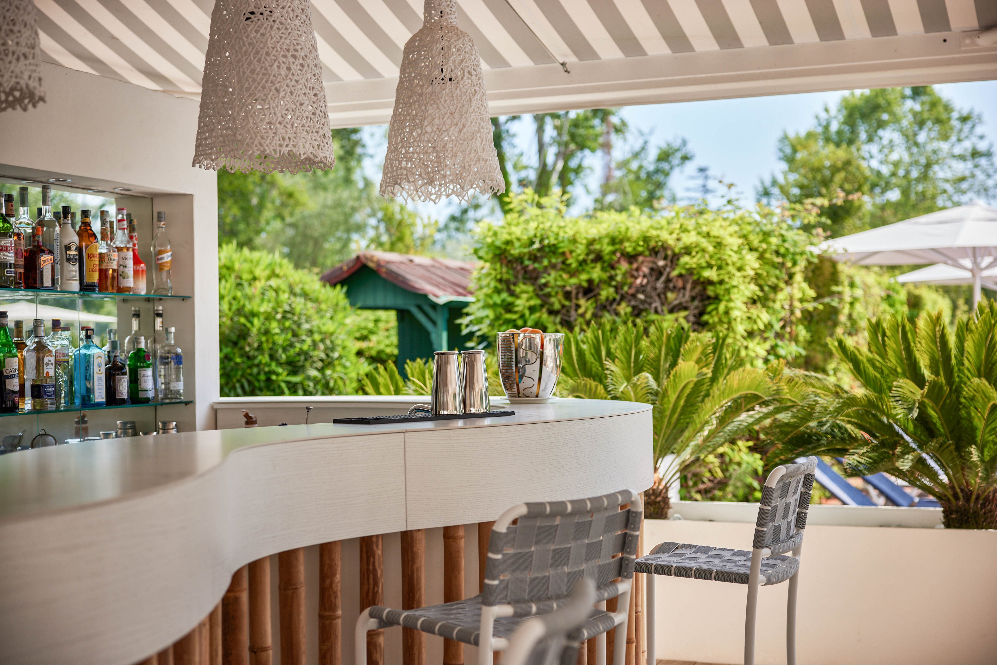 A stylish outdoor bar features a curved white counter with two woven chairs in the foreground. The bar is stocked with various bottles and glassware, set against a lush green garden backdrop. Overhead, woven pendant lights and a striped canopy add a relaxed, summery vibe. The setting suggests a tranquil, resort-like atmosphere ideal for leisure and socializing.