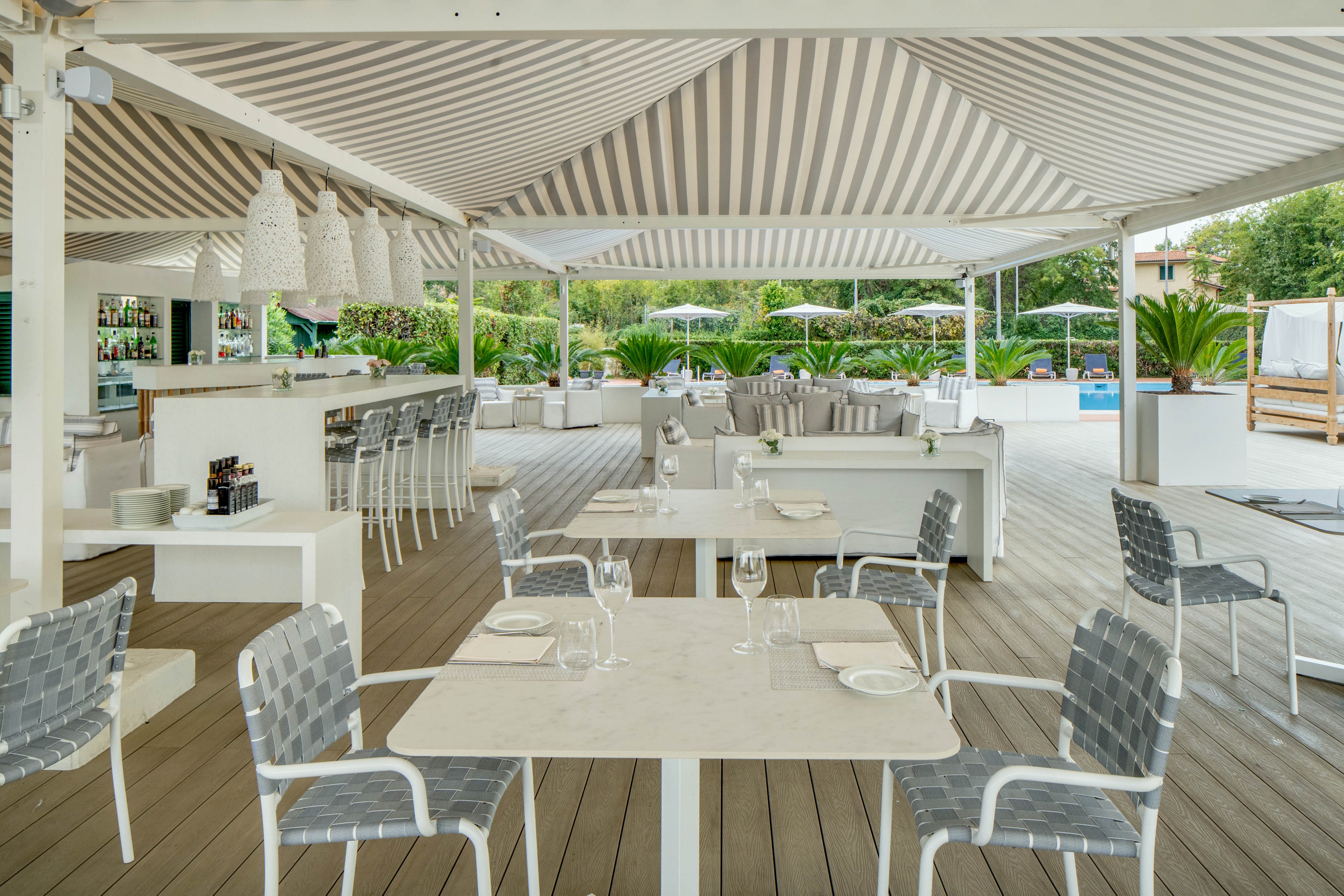 A contemporary outdoor restaurant features white tables and woven chairs under a striped canopy. The setting includes a stylish bar area, lounge sofas, and a view of a pool surrounded by greenery. The atmosphere is bright and inviting, with neutral tones and natural light. No visible people are present, emphasizing a serene and relaxed mood. Table settings include glassware and plates, ready for guests.