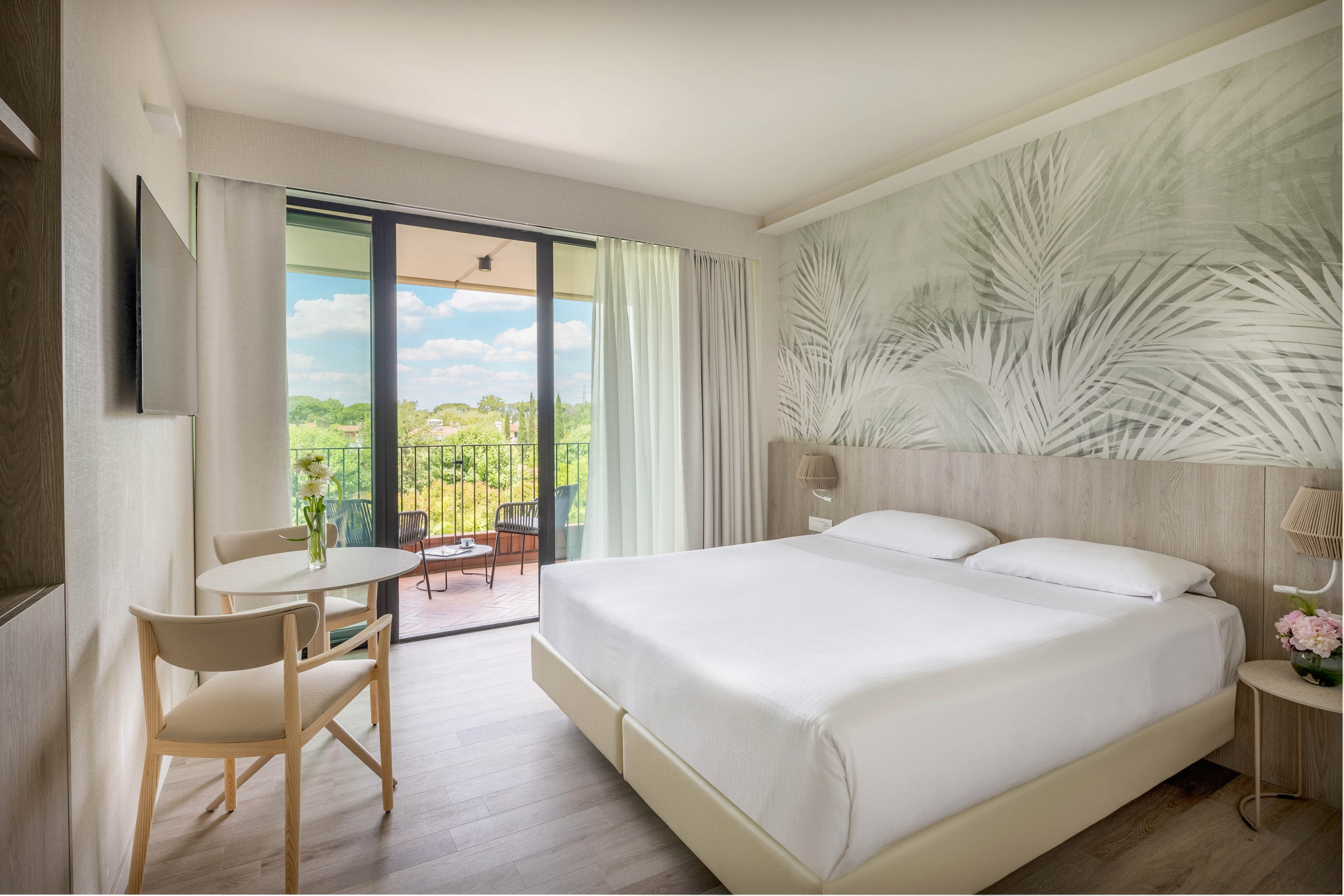 A bright, contemporary hotel room features a large double bed, minimalist furniture, and a wall mural with botanical patterns. The room opens to a balcony with outdoor seating, overlooking a lush green landscape under a blue sky. Natural light fills the space, creating a calm and inviting atmosphere, with neutral tones and soft accents throughout.
