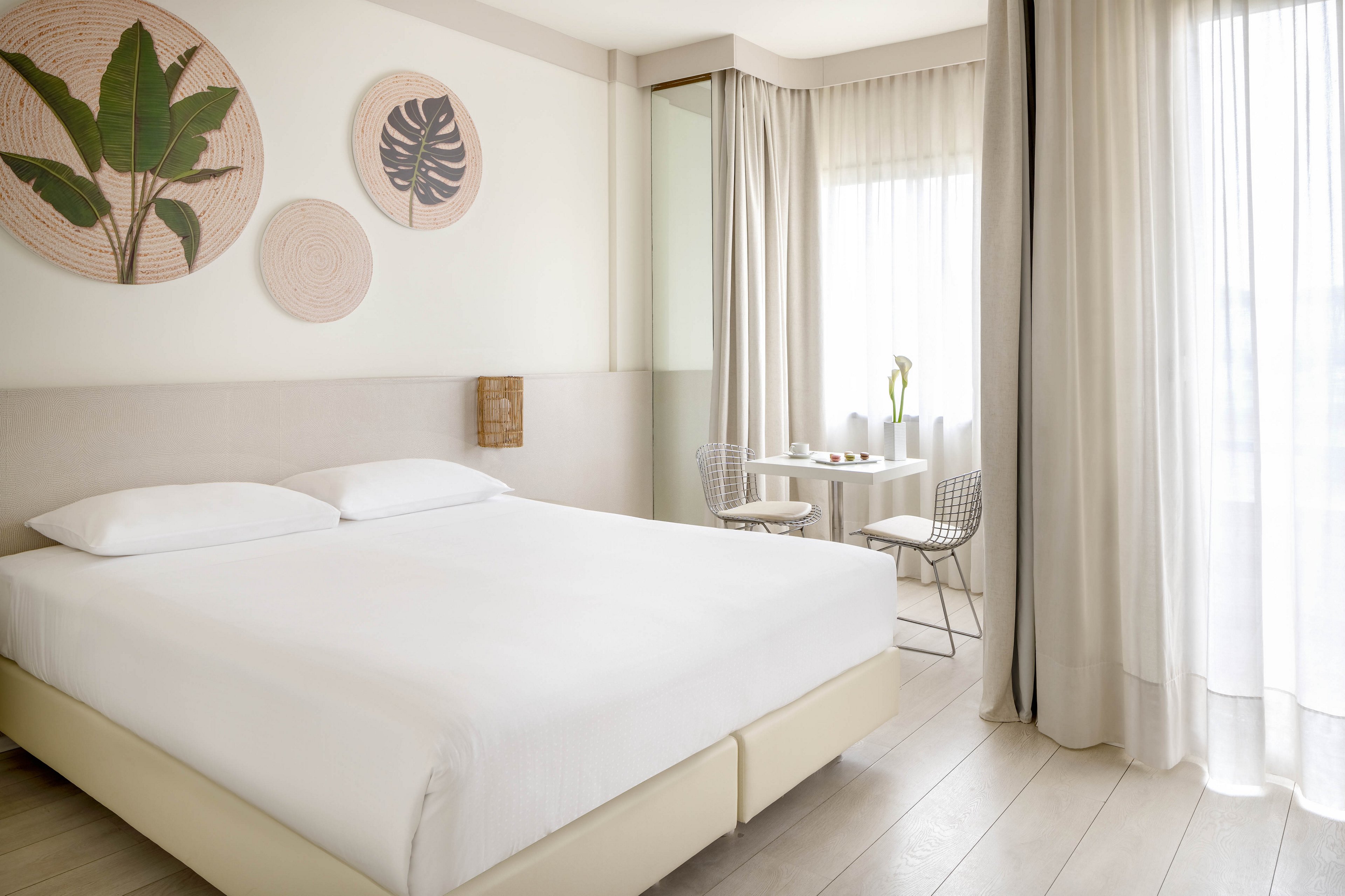 A spacious hotel room features a large bed with crisp white linens and neutral decor. The room is filled with natural light from sheer curtains covering tall windows. Wall art with botanical designs adds a touch of nature, while a small table and two chairs sit by the window. The overall atmosphere is calm, airy, and modern.