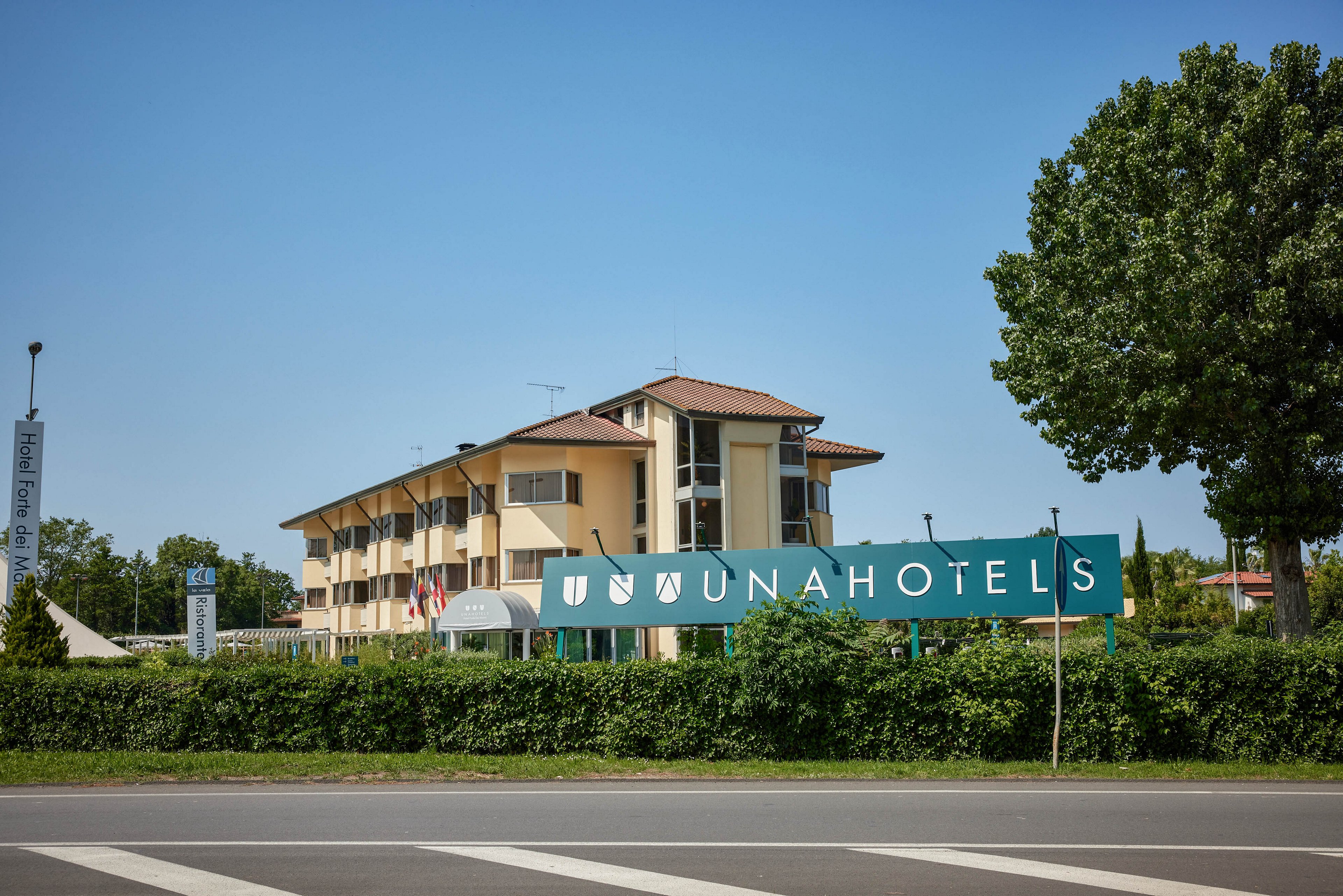 A large UNA Hotels building is prominently displayed behind a green sign with the UNA Hotels logo and text. The hotel is set in a suburban or semi-rural area with a clear blue sky and lush greenery. Multiple flags are visible near the entrance, and the scene is captured from across a road with visible crosswalk lines. The overall mood is bright and welcoming.