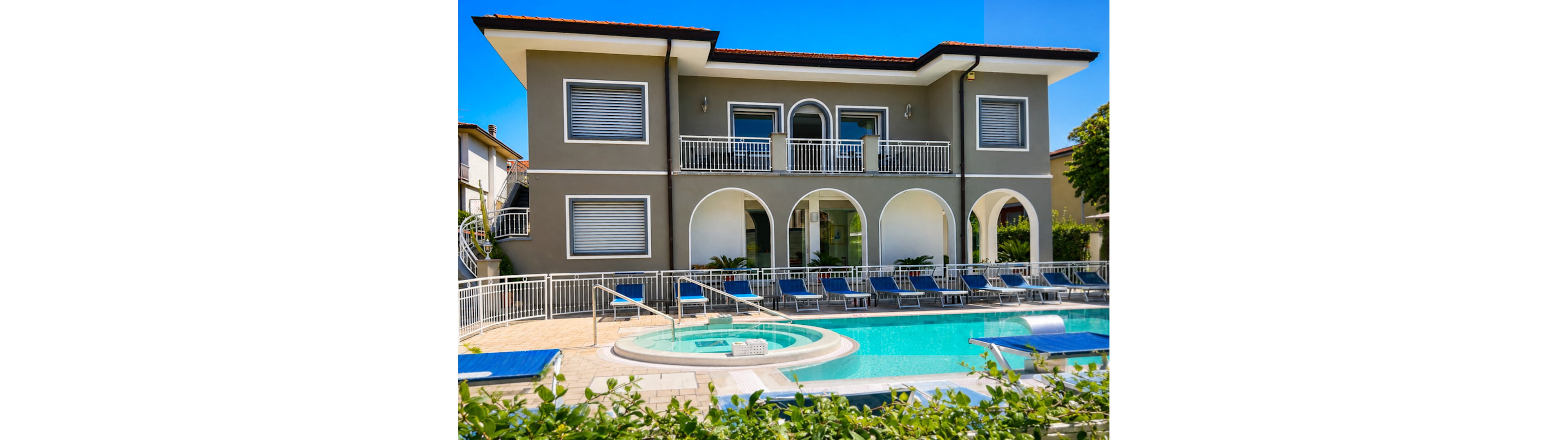 A contemporary two-story villa features a row of arched openings facing a bright blue swimming pool. Sun loungers and umbrellas line the poolside terrace, suggesting a relaxing vacation setting. The building has clean architectural lines, white railings, and large shuttered windows. Lush greenery in the foreground and a clear blue sky enhance the summery, resort-like atmosphere.