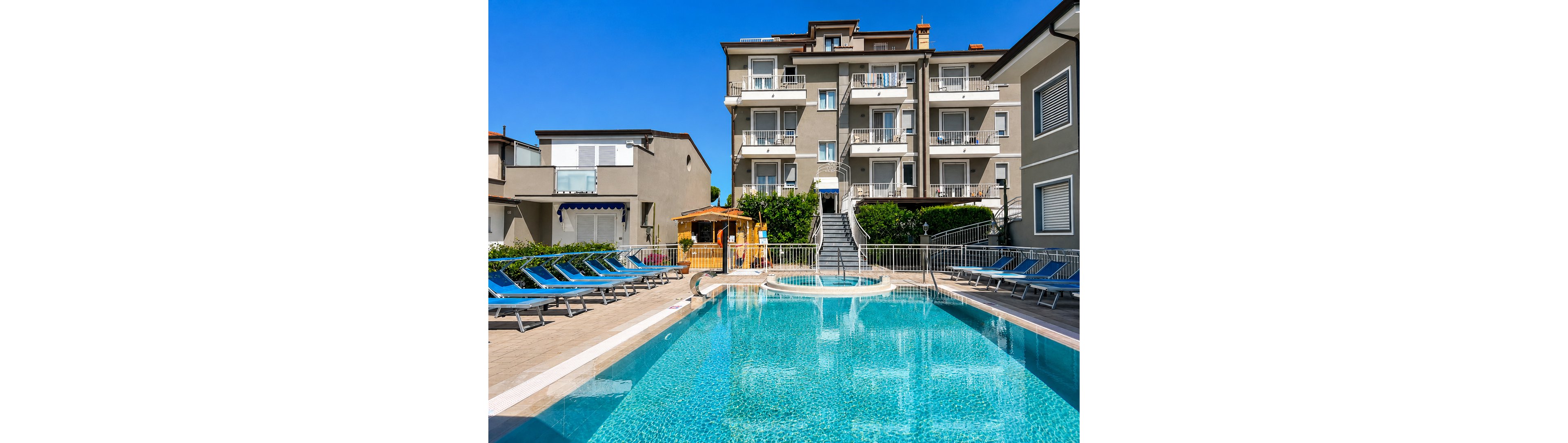 A clear blue swimming pool sits in the center of a hotel courtyard under a bright, cloudless sky. Multiple lounge chairs line both sides of the pool, suggesting a relaxing sunbathing area. A small wooden kiosk or bar stands near the stairs that lead up to the multi-story residential-style building in the background. The overall scene conveys a clean, inviting vacation atmosphere with modern architecture and well-kept surroundings.