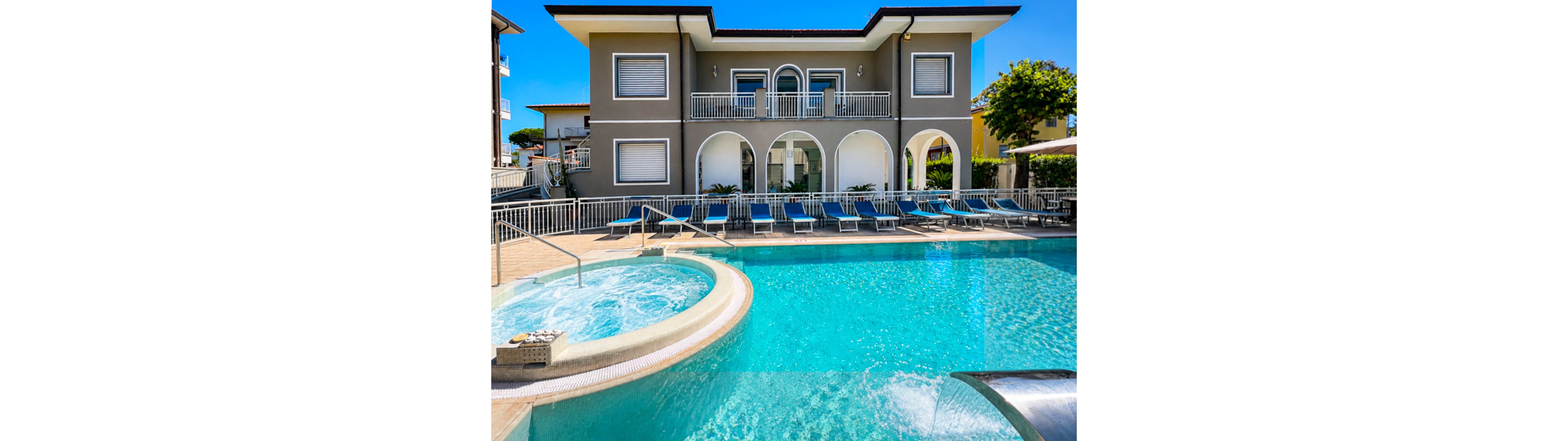 A contemporary two-story villa features a bright blue outdoor swimming pool in the foreground. Multiple sun loungers line the poolside terrace, suggesting a relaxing vacation setting. An attached circular spa pool with metal handrails and a curved waterfall spout adds a luxurious touch. The building’s arched ground-floor openings, balconies, and clean architectural lines stand out under a clear sunny sky.