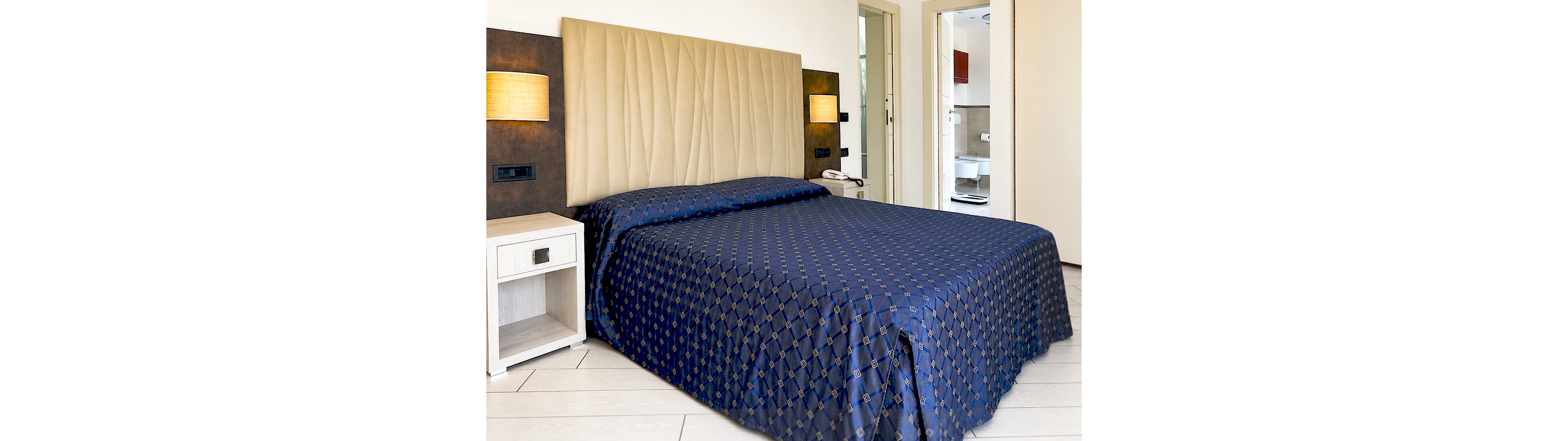 A neatly made double bed with a patterned blue bedspread is the focal point of a modern hotel room. The bed features a tall, upholstered beige headboard against a dark accent wall with built-in lighting. A white bedside table with a drawer and open shelf stands next to the bed. In the background, an open doorway reveals a bright bathroom area, adding depth to the interior scene. The overall space appears clean, contemporary, and inviting for guests.