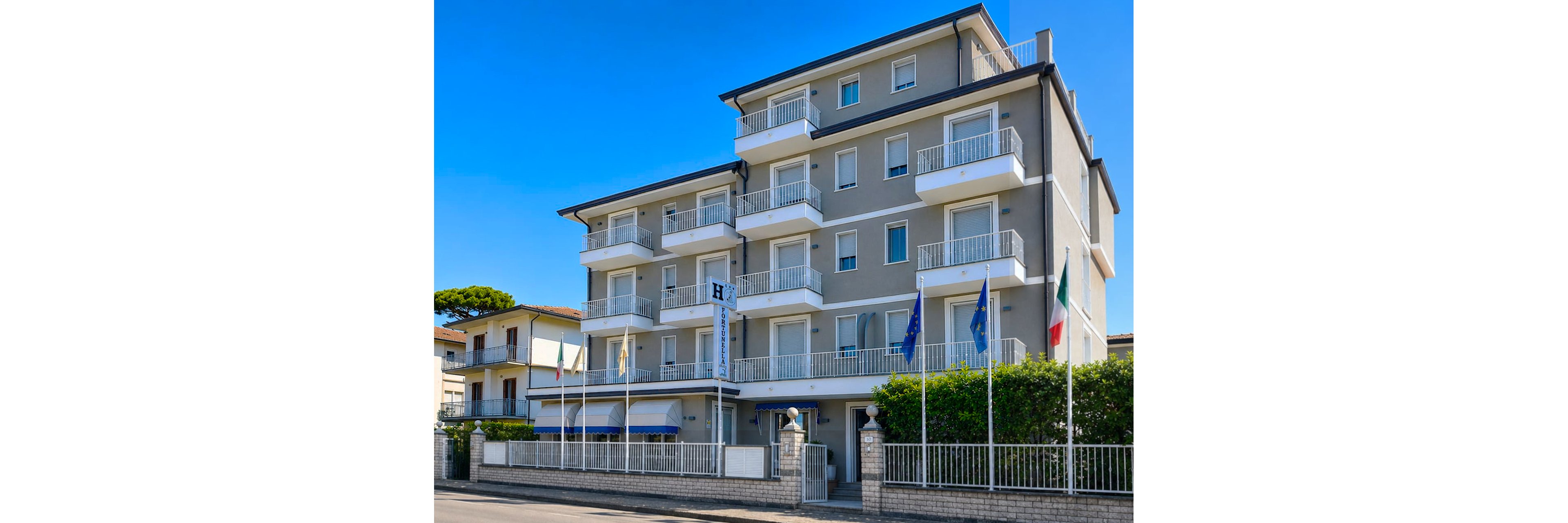 A contemporary four-story hotel stands along a quiet street, featuring multiple balconies with white railings and large windows. The facade is light gray with a darker roofline, and a fenced hedge borders the sidewalk in front. Several flags are displayed on tall poles near the entrance, adding color to the scene. Neighboring residential buildings and a clear blue sky complete the bright daytime setting.