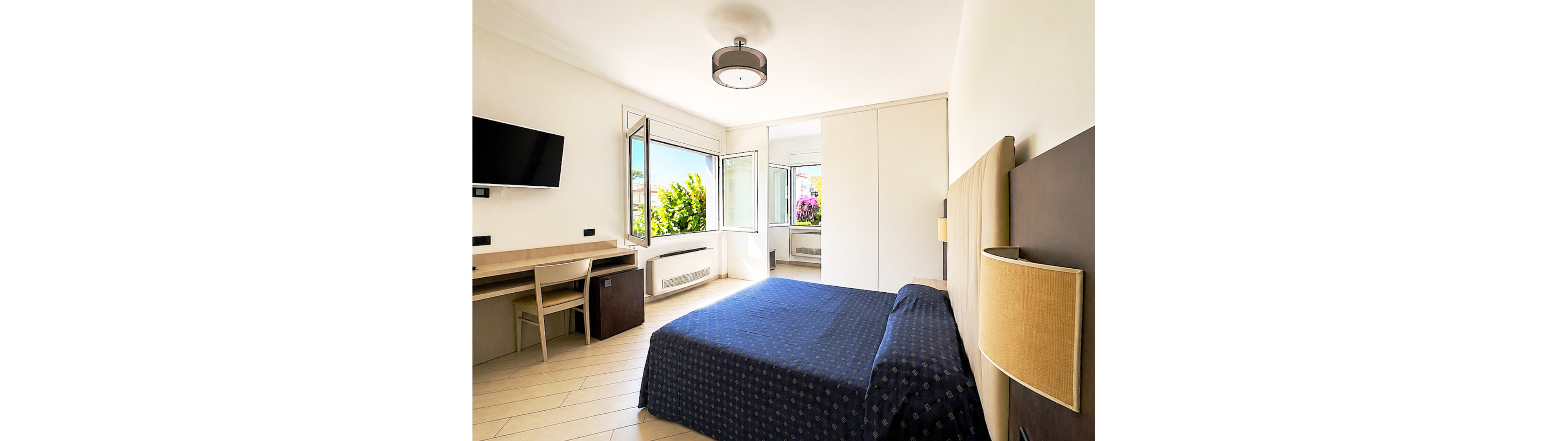 A modern hotel-style bedroom features a neatly made double bed with a dark blue patterned bedspread. Large windows are open, letting in natural light and revealing greenery and flowering plants outside. A wall-mounted flat-screen TV faces the bed, and a simple wooden desk with chair sits along one wall. The room is decorated in neutral tones with minimal furnishings, creating a clean and airy atmosphere.