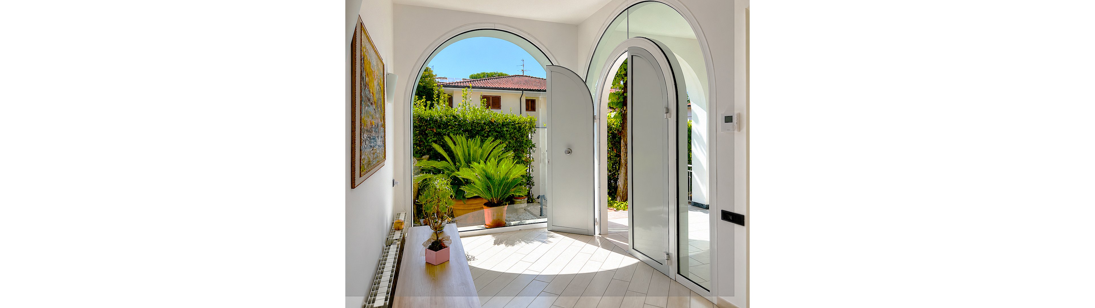 A sunlit home entryway features large white arched doors opening to a lush garden. Light wood or tile flooring reflects the natural light streaming in from the doorway and windows. Potted green plants sit on a narrow console table and on the floor, adding a fresh, natural accent to the interior. A framed wall painting and outdoor view of neighboring houses complete the welcoming residential scene.
