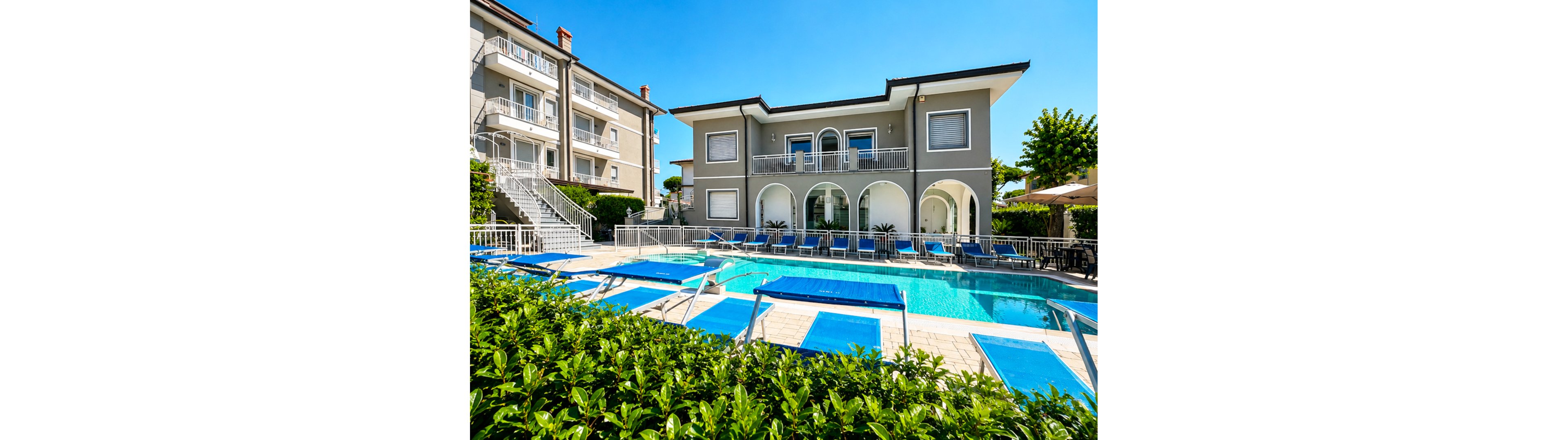 A contemporary two-story villa features a bright blue outdoor swimming pool surrounded by sun loungers. The building has arched ground-floor windows, balconies with white railings, and a clean, neutral facade. Neighboring apartments and a staircase are visible to the left, while greenery and poolside umbrellas frame the scene. The clear blue sky and vivid colors create a sunny, inviting holiday atmosphere.