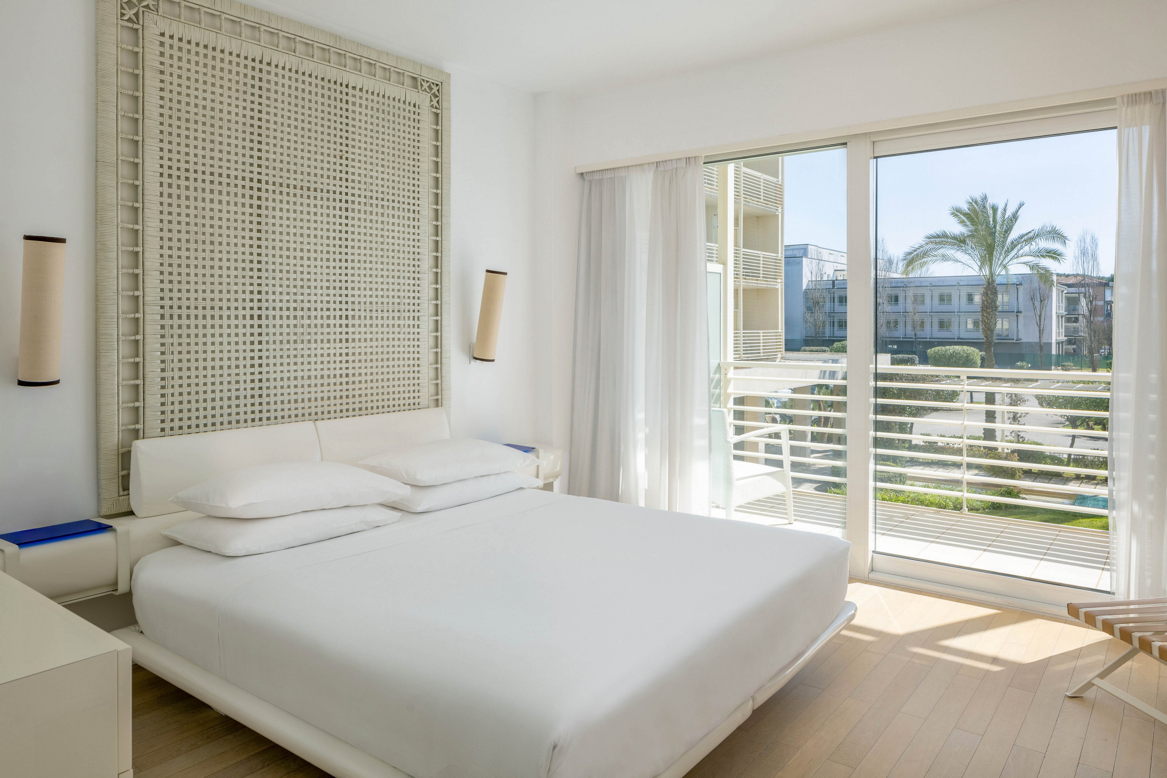 A bright, modern hotel bedroom features a large white bed and minimalist decor. The room opens to a balcony with glass doors, offering a view of palm trees and a sunny outdoor area. Light wood flooring and white walls create an airy, serene atmosphere. No visible text or numbers are present in the image.