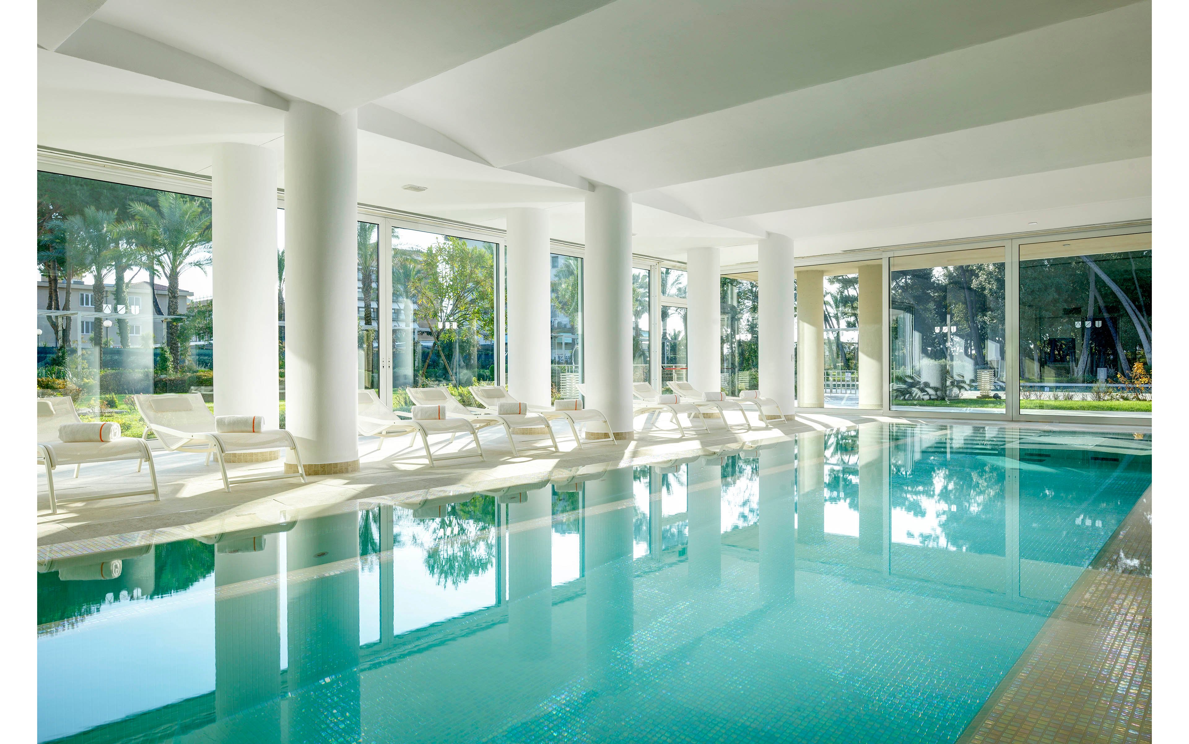 A spacious indoor swimming pool is surrounded by white lounge chairs and large floor-to-ceiling windows. Natural light fills the area, creating a serene and inviting atmosphere. The pool reflects the modern architecture and lush greenery visible outside. The setting is calm, with no people present, emphasizing relaxation and tranquility.