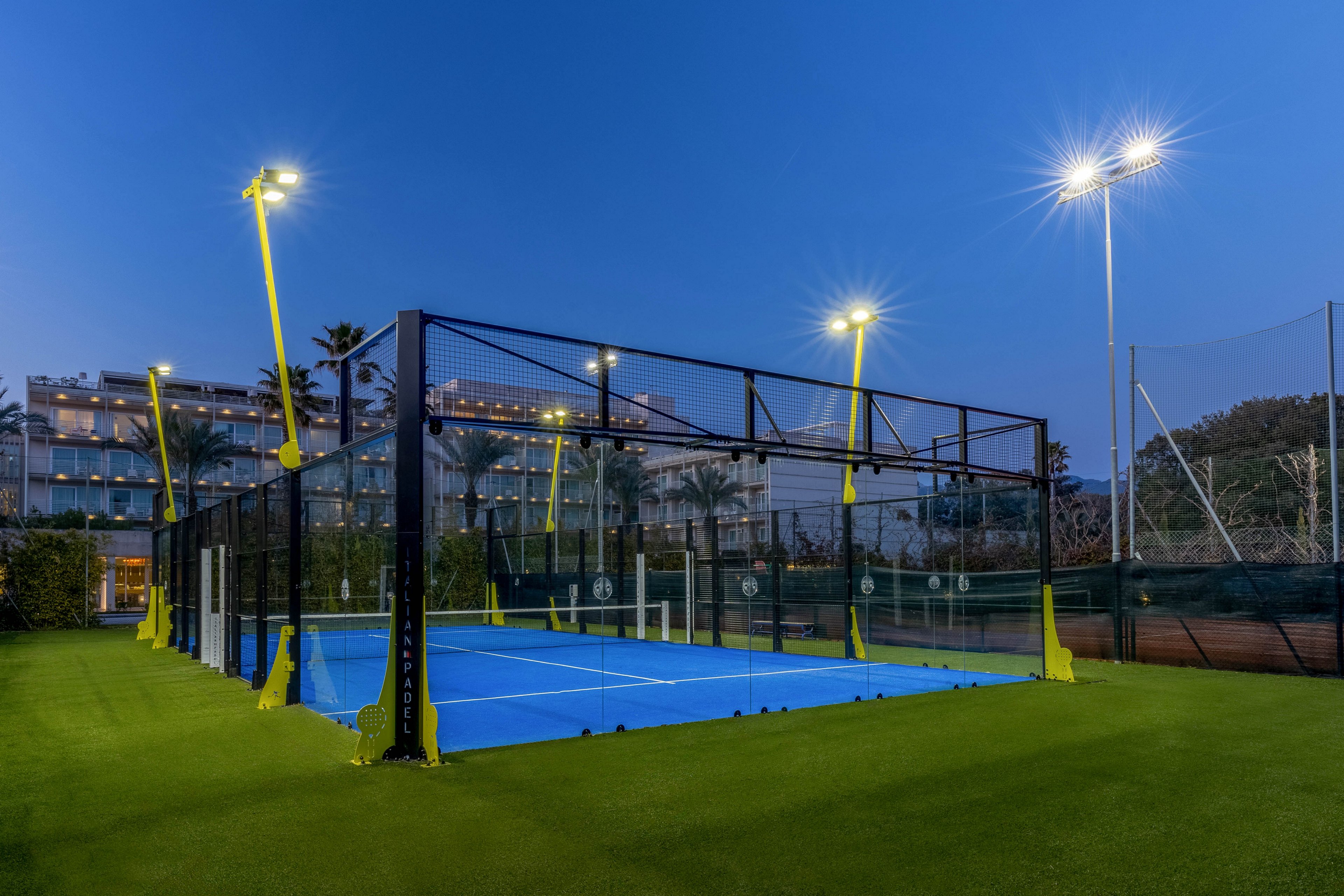 A modern outdoor padel court is brightly lit under tall floodlights during the early evening. The blue playing surface contrasts with the green artificial turf surrounding the court. In the background, a multi-story building and palm trees are visible, adding to the urban sports setting. The sky is clear, transitioning from daylight to night.