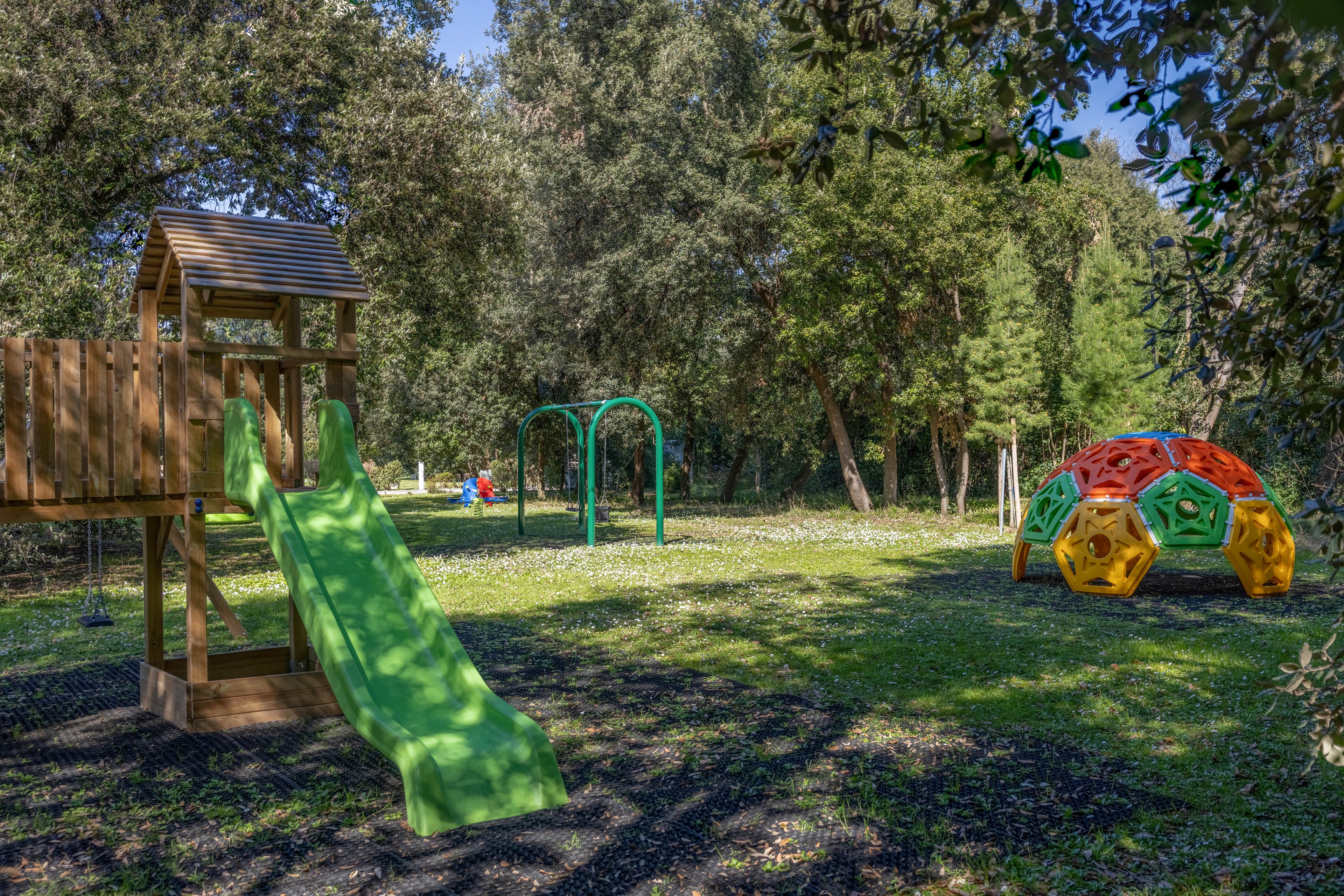 A vibrant outdoor playground features a green slide attached to a wooden structure, a set of green swings, and a colorful geometric climbing dome. The play area is surrounded by lush trees and grass, creating a natural and inviting setting. Sunlight filters through the foliage, casting playful shadows on the ground. No visible text or numbers are present in the image.