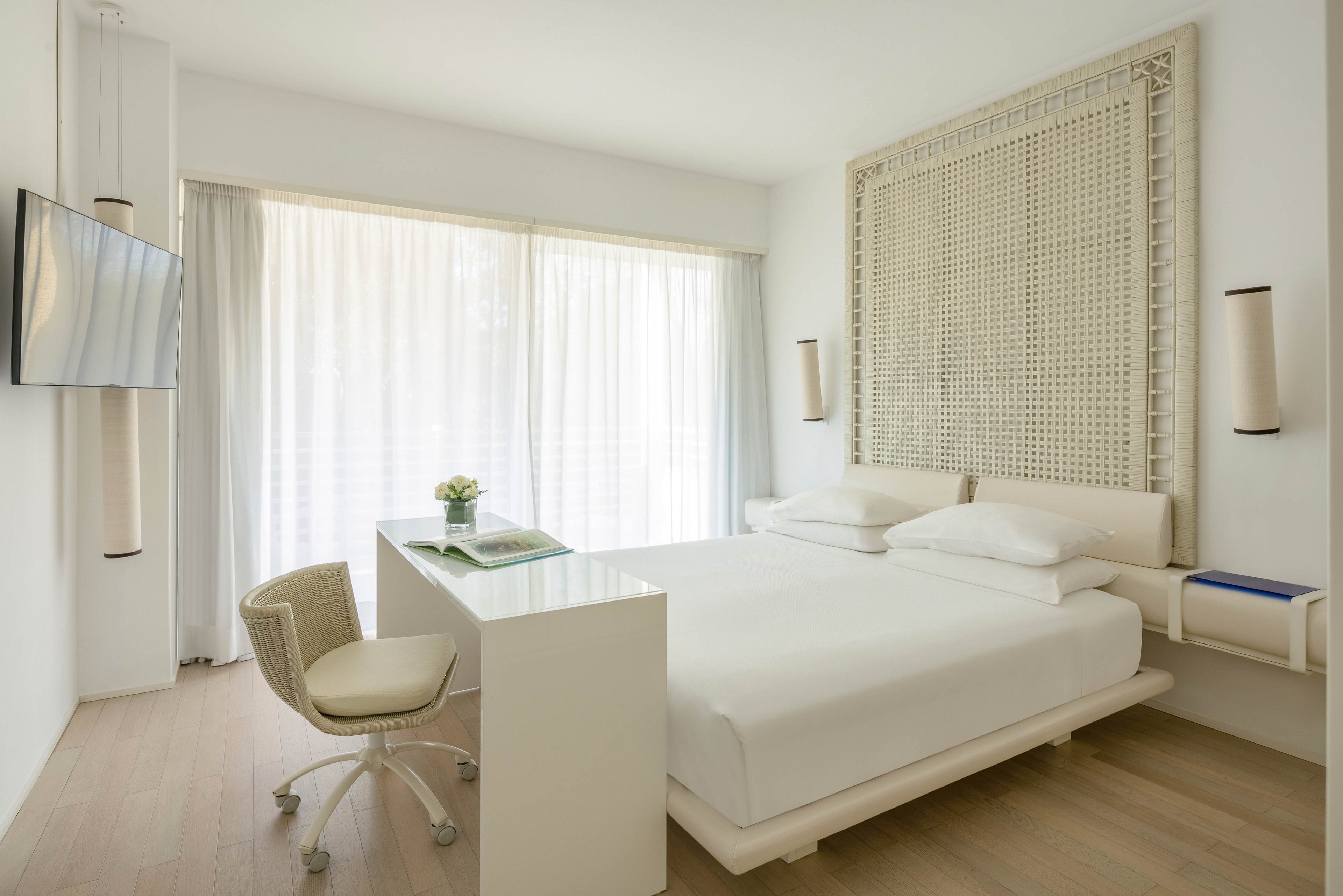 A bright, minimalist hotel room features a large bed with crisp white linens and a decorative headboard. The space includes a sleek white desk and chair, a wall-mounted television, and floor-to-ceiling windows with sheer curtains letting in natural light. The room is styled in a neutral palette with light wood flooring and subtle decorative accents.
