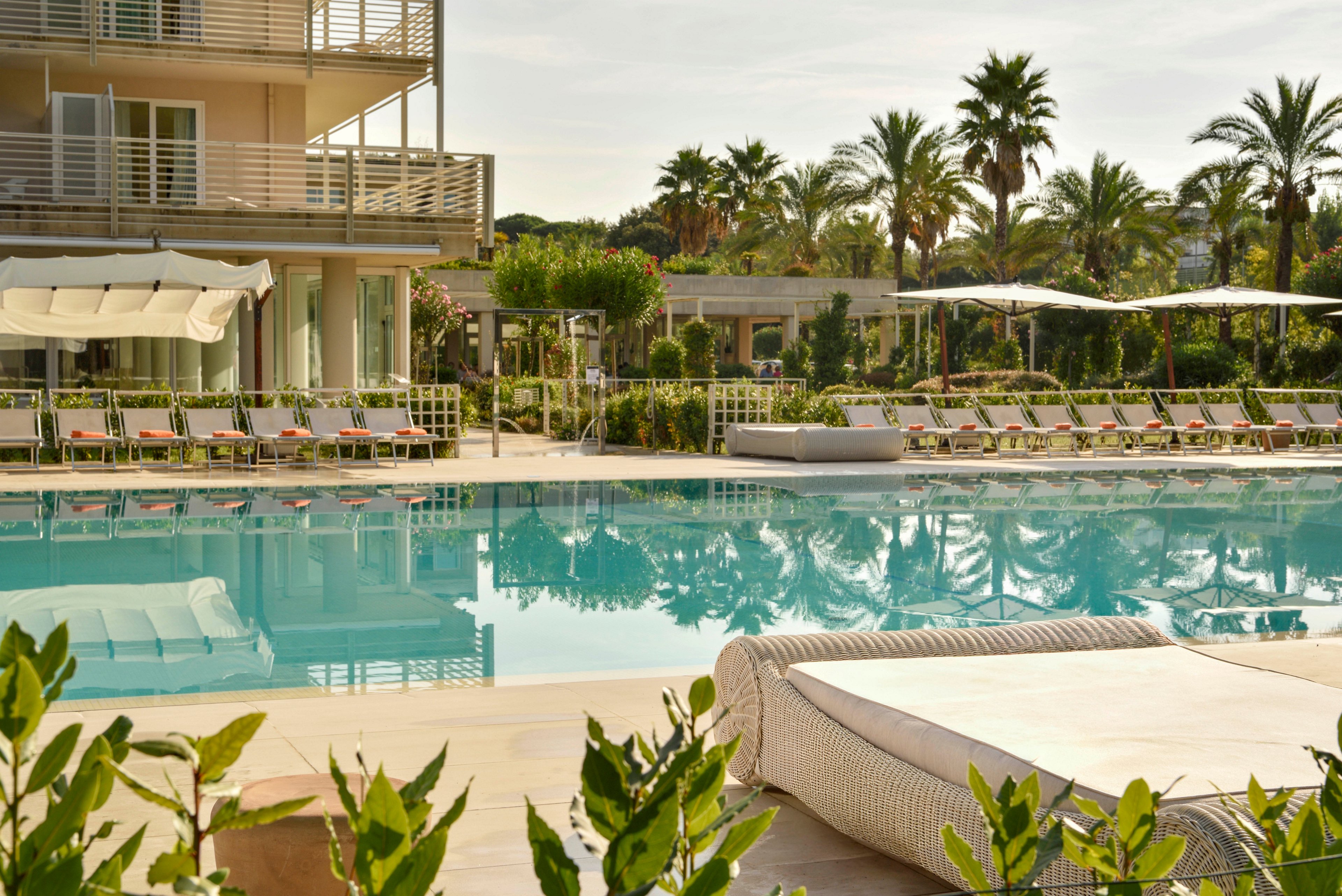 A woman in a floral dress and sunhat lounges on a poolside chair, holding a pink beverage. The setting is a modern resort with palm trees, sun loungers, and a large outdoor swimming pool. The atmosphere is tranquil and summery, with warm sunlight and lush greenery. The scene evokes a sense of leisure and vacation relaxation.