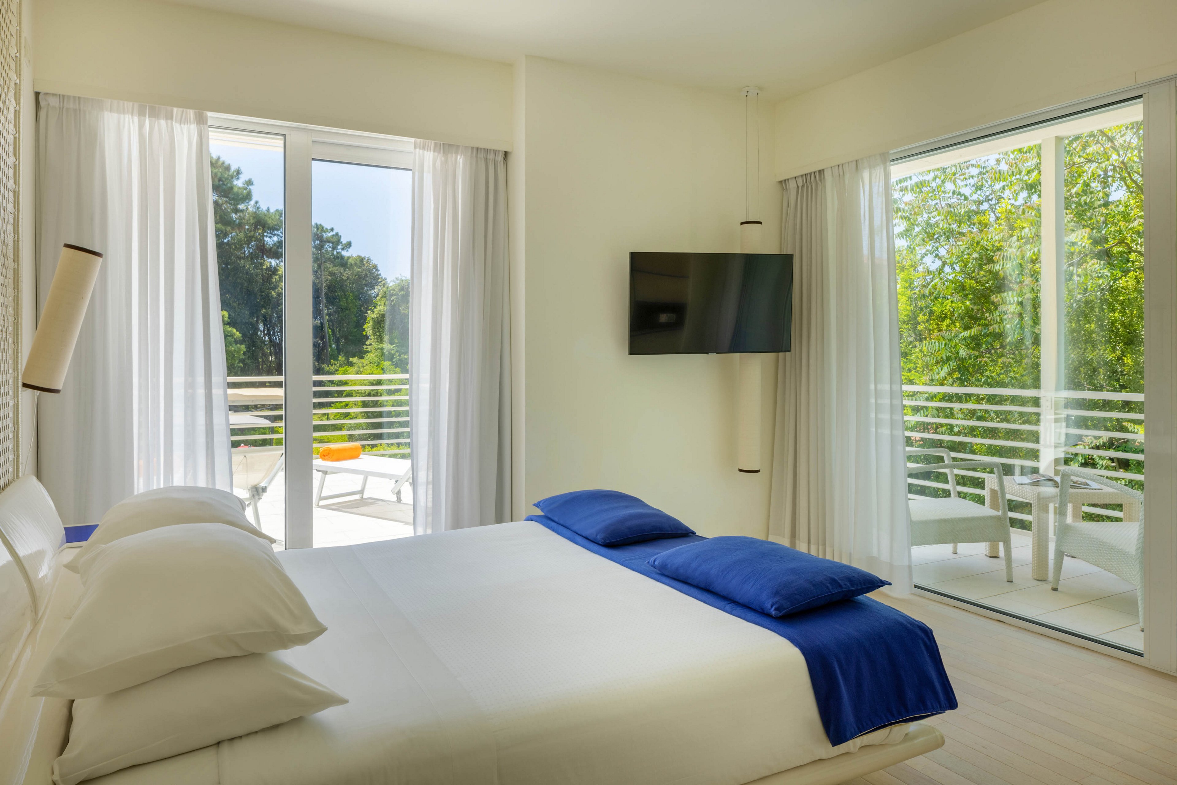 A bright, contemporary hotel room features a large bed with crisp white linens and blue pillows. The space is illuminated by natural light from two large glass doors leading to a balcony with outdoor seating. Minimalist decor and a wall-mounted television enhance the clean, modern aesthetic. Lush greenery is visible outside, creating a tranquil atmosphere. No visible text or numbers are present in the image.