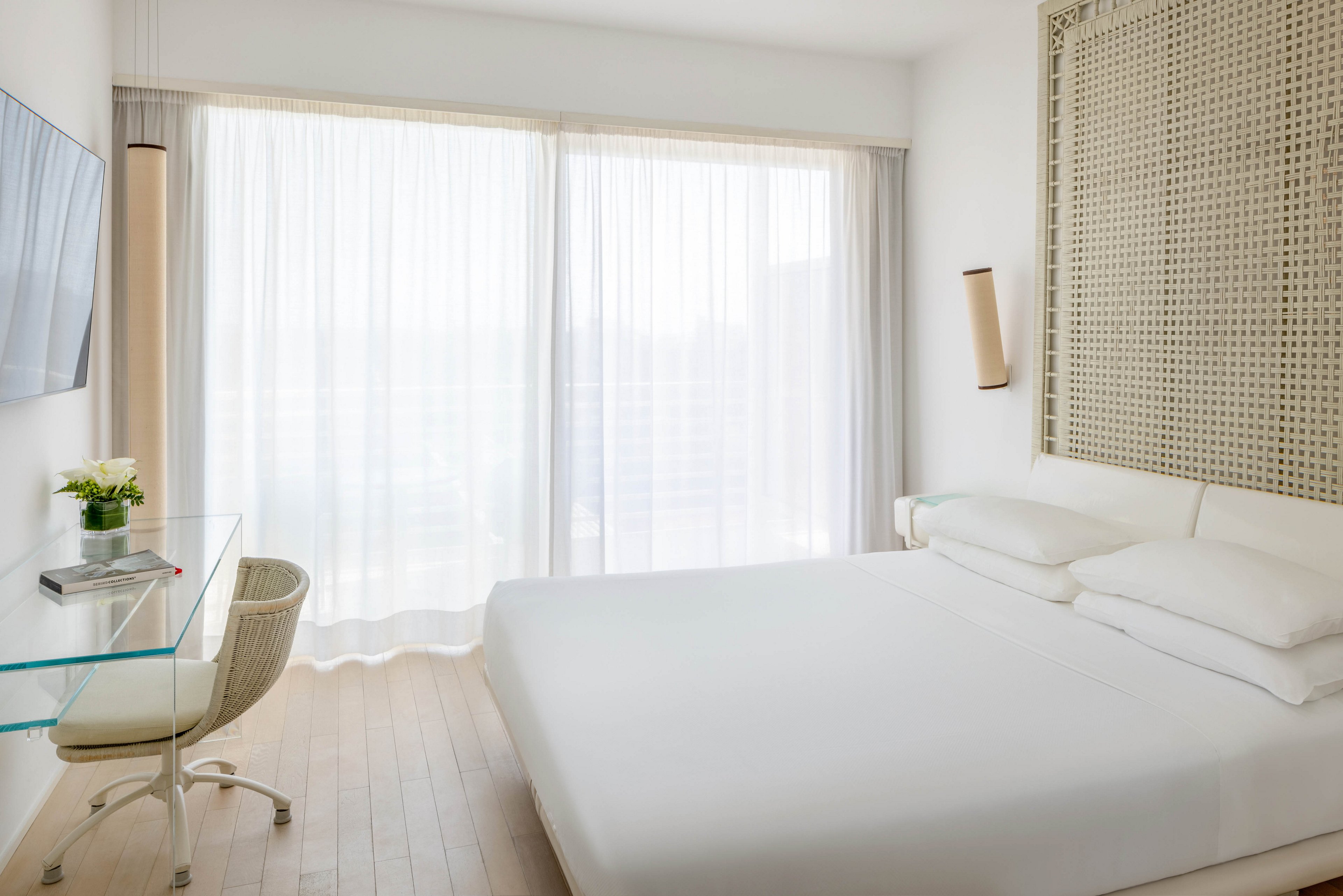 A modern hotel room features a large bed with white linens, a glass desk with a chair, and floor-to-ceiling windows covered by sheer curtains. The space is decorated in a minimalist style with light wood flooring and neutral tones. A small vase with flowers sits on the desk, and the room is filled with natural daylight, creating an airy and serene atmosphere.