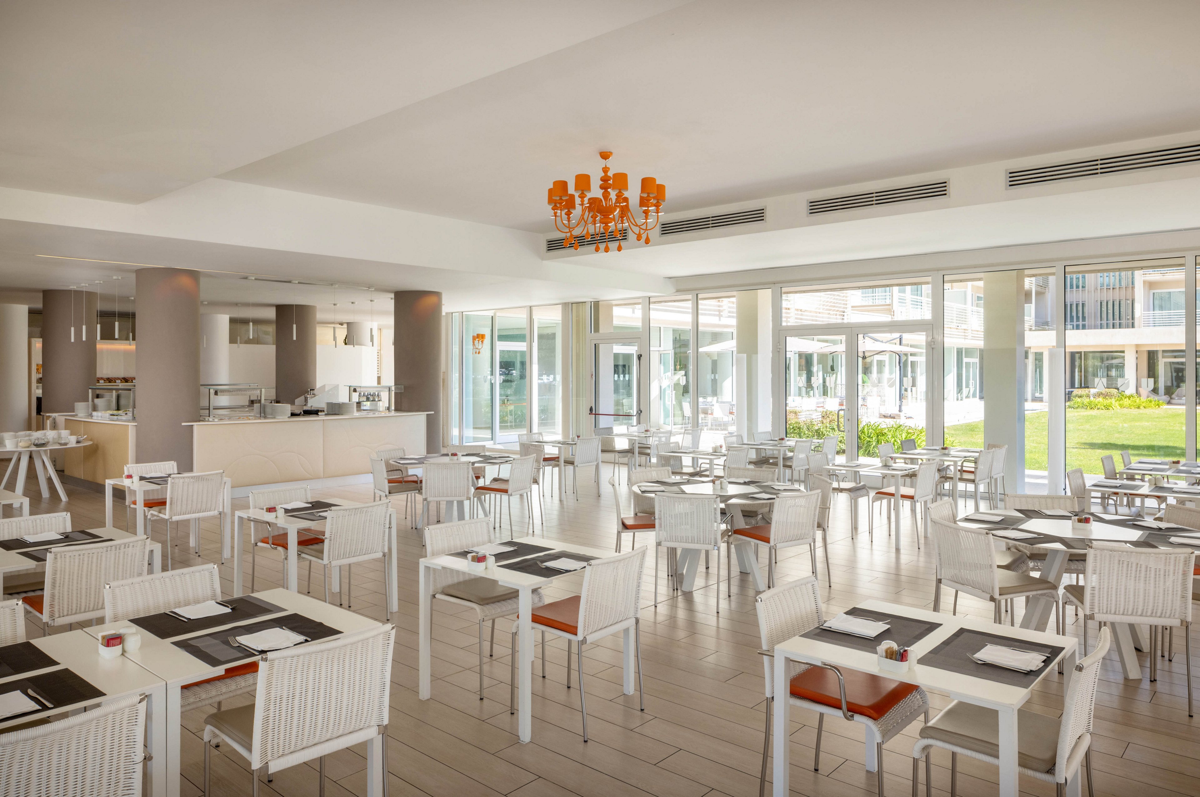 A spacious, contemporary restaurant dining area features white tables and chairs arranged neatly across a tiled floor. Large windows allow natural light to fill the room, offering views of a green outdoor space. The setting is accented by a bold orange chandelier, creating a fresh and inviting atmosphere. Place settings are minimal, with black placemats and white dishware. No people are present, emphasizing the clean and organized environment.