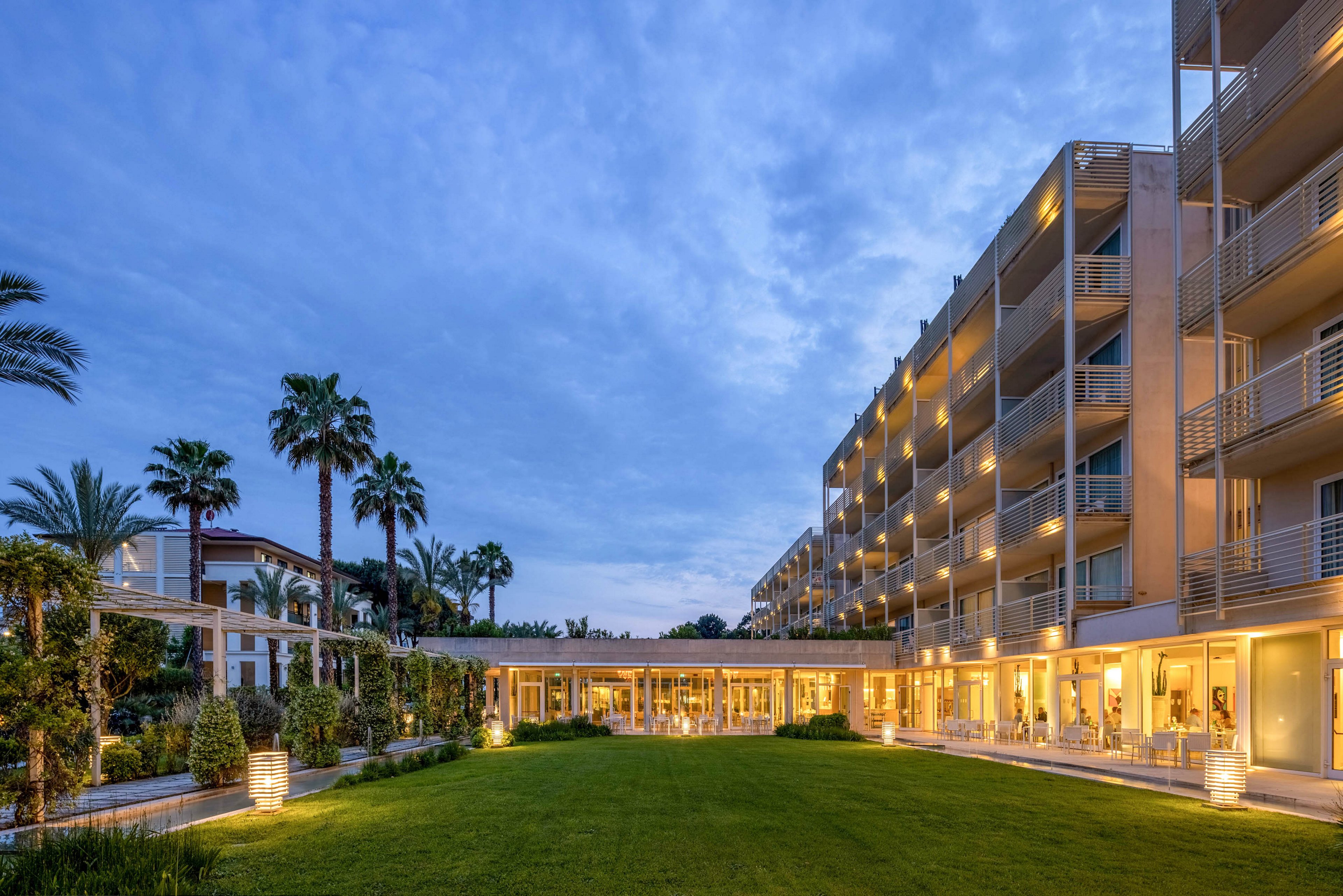 A contemporary multi-story hotel building is shown at twilight, with warm lights glowing from balconies and windows. The scene features a manicured lawn and palm trees, creating a tranquil and inviting atmosphere. The sky is a deep blue, enhancing the evening ambiance. No visible text or numbers are present in the image.