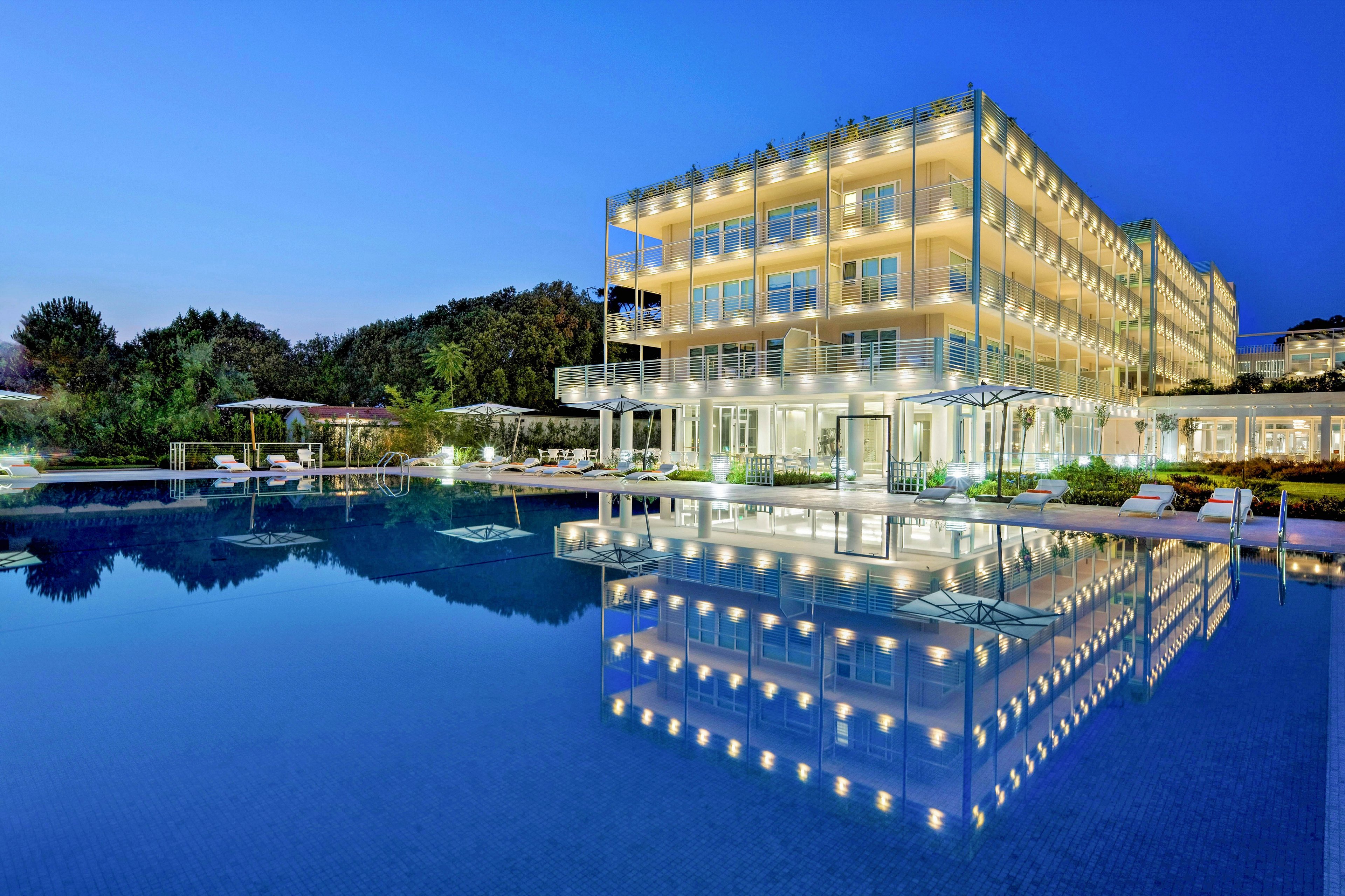 A contemporary multi-story hotel is beautifully lit up in the evening, with its lights reflecting in a large, tranquil outdoor swimming pool. The setting is serene, featuring lounge chairs and umbrellas around the pool area, suggesting a relaxing atmosphere. The sky is a deep blue, enhancing the luxurious and inviting mood of the scene. No visible text or numbers are present in the image.