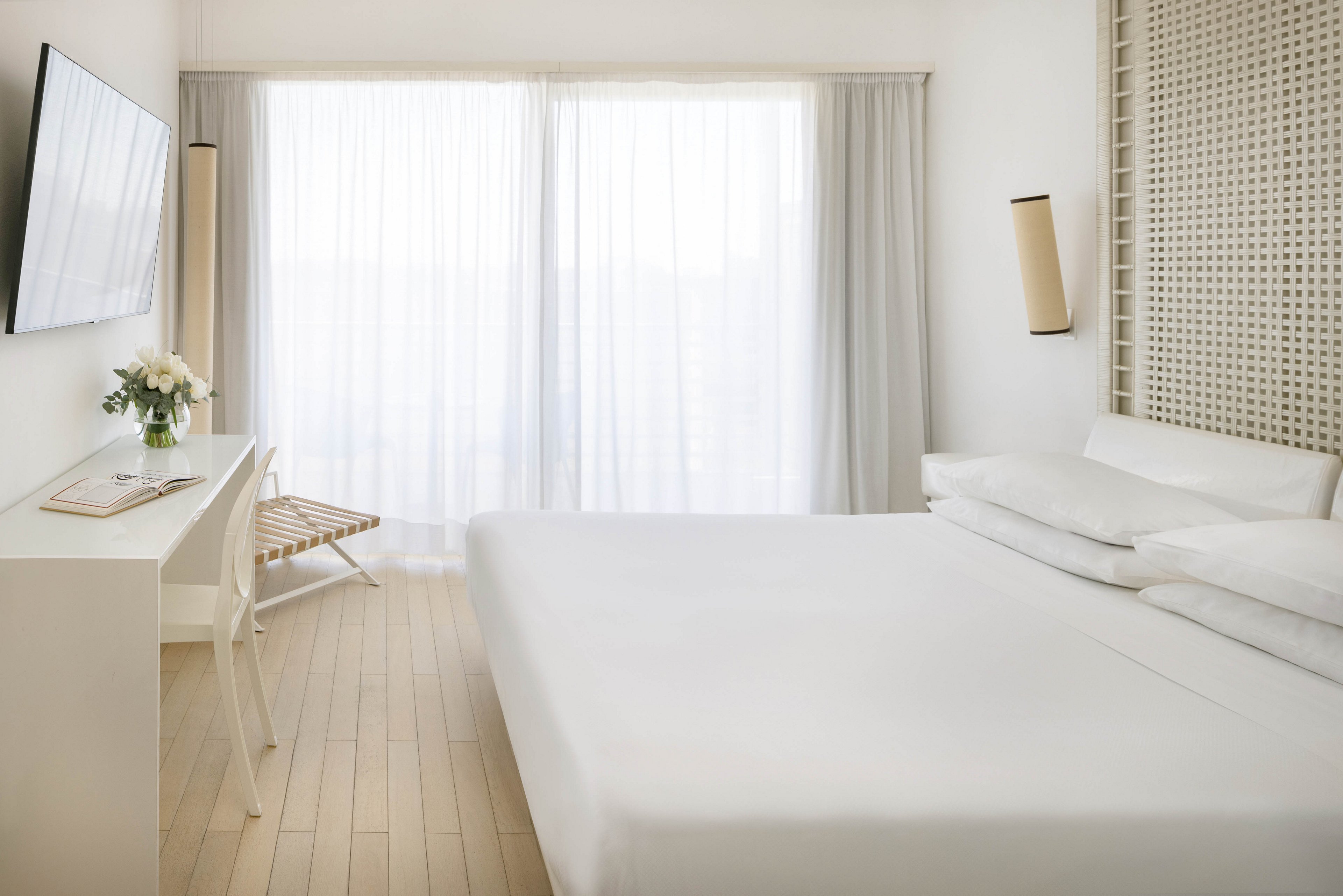 A bright, minimalist hotel room features a large bed with crisp white linens and a sleek desk area. Natural light pours in through sheer curtains covering a wide window, creating an airy and serene atmosphere. The room is decorated in neutral tones with light wood flooring and simple, modern furnishings. No visible text or numbers are present in the image.