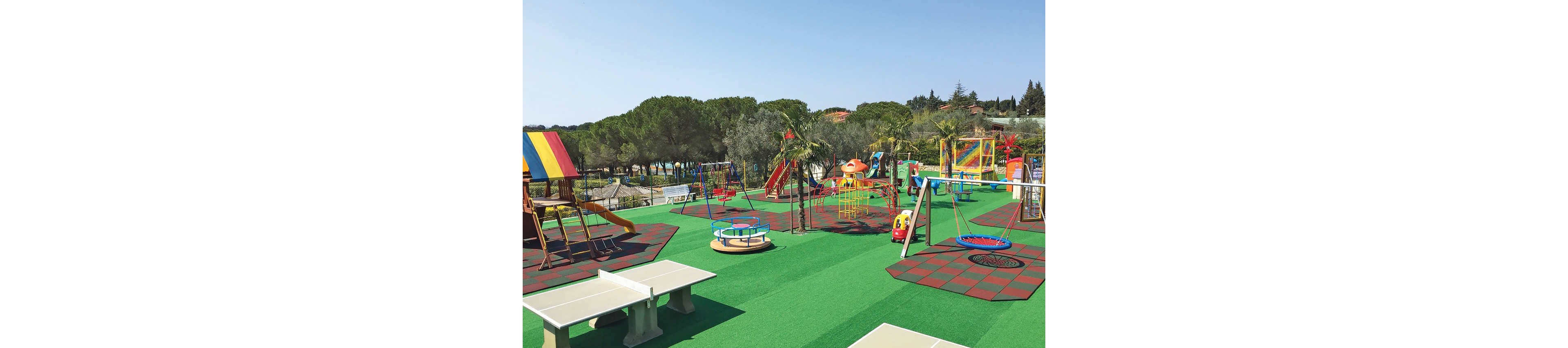 A bright outdoor playground features multiple play structures on a green rubber surface. Swings, slides, climbing frames, and a merry-go-round are arranged across the area, with some sections set on red and black safety tiles. The playground is surrounded by trees and greenery, suggesting a park or recreational area. No people are present, highlighting the equipment and colorful design.