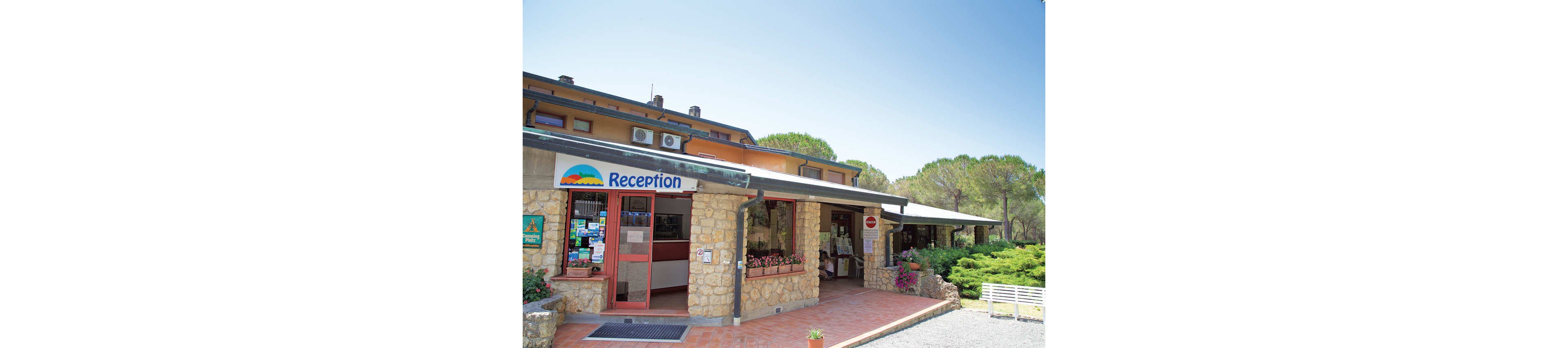 A stone and brick reception building stands at the entrance of a campground or holiday park. The sign above the door clearly reads "Reception" with a colorful landscape graphic. Large windows display brochures and information, while potted flowers decorate the doorway. Surrounding the structure are green shrubs, tall trees, and a gravel path under a bright blue sky.