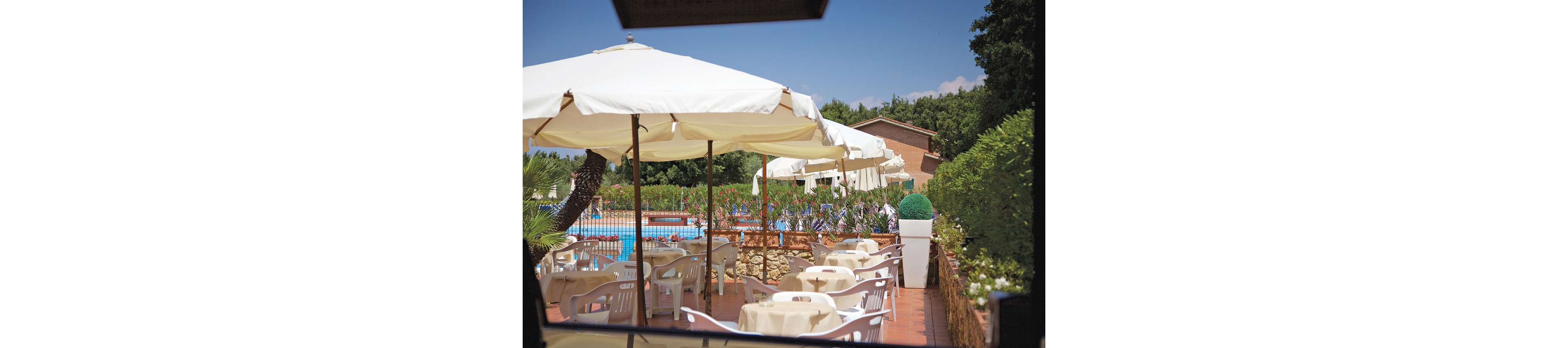 An outdoor café area features several white tables and chairs arranged under large cream-colored umbrellas. The seating overlooks a bright blue swimming pool surrounded by a low stone wall and lush greenery. In the background, a house-like building and tall trees frame the relaxed leisure setting. The scene suggests a sunny, tranquil day at a resort or holiday property.