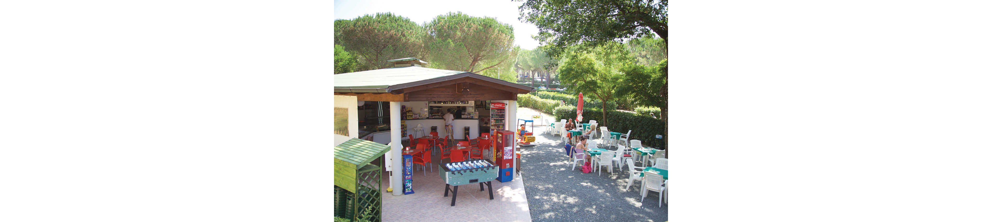 An open-air café with a wooden roof features bright orange chairs and tables around a central bar counter. A foosball table and vending machines stand near the entrance, adding to the casual atmosphere. Outside, white plastic chairs and tables are arranged on a gravel terrace under large trees, where a few people relax. The surrounding greenery and tall pines create a shaded, holiday-like setting on a sunny day.