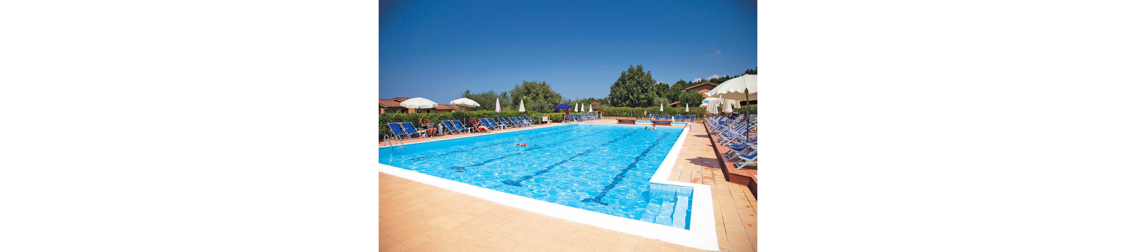 A large rectangular outdoor swimming pool glistens under a clear blue sky. Sun loungers with blue fabric line both sides of the pool, many shaded by white umbrellas. A few people relax on the chairs while one swimmer enjoys the water. Trees and low buildings in the background create a peaceful resort atmosphere.