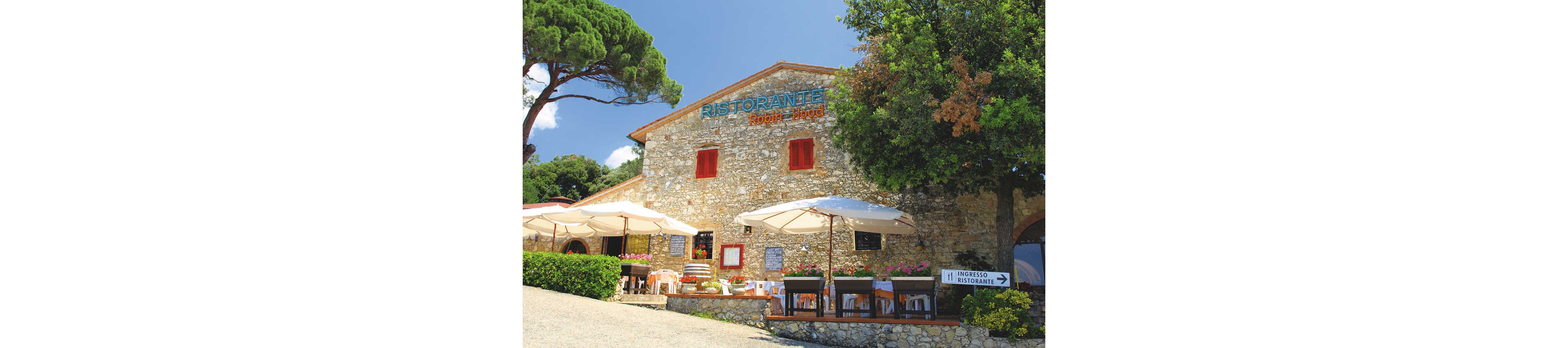 A rustic stone ristorante with bright red window frames features an inviting outdoor dining terrace. Tables and chairs are arranged under large white umbrellas, surrounded by flowers and greenery. The building is set on a gentle slope with trees framing the scene under a clear sky. A sign reading "INGRESSO RISTORANTE" directs guests toward the entrance, enhancing the welcoming atmosphere.